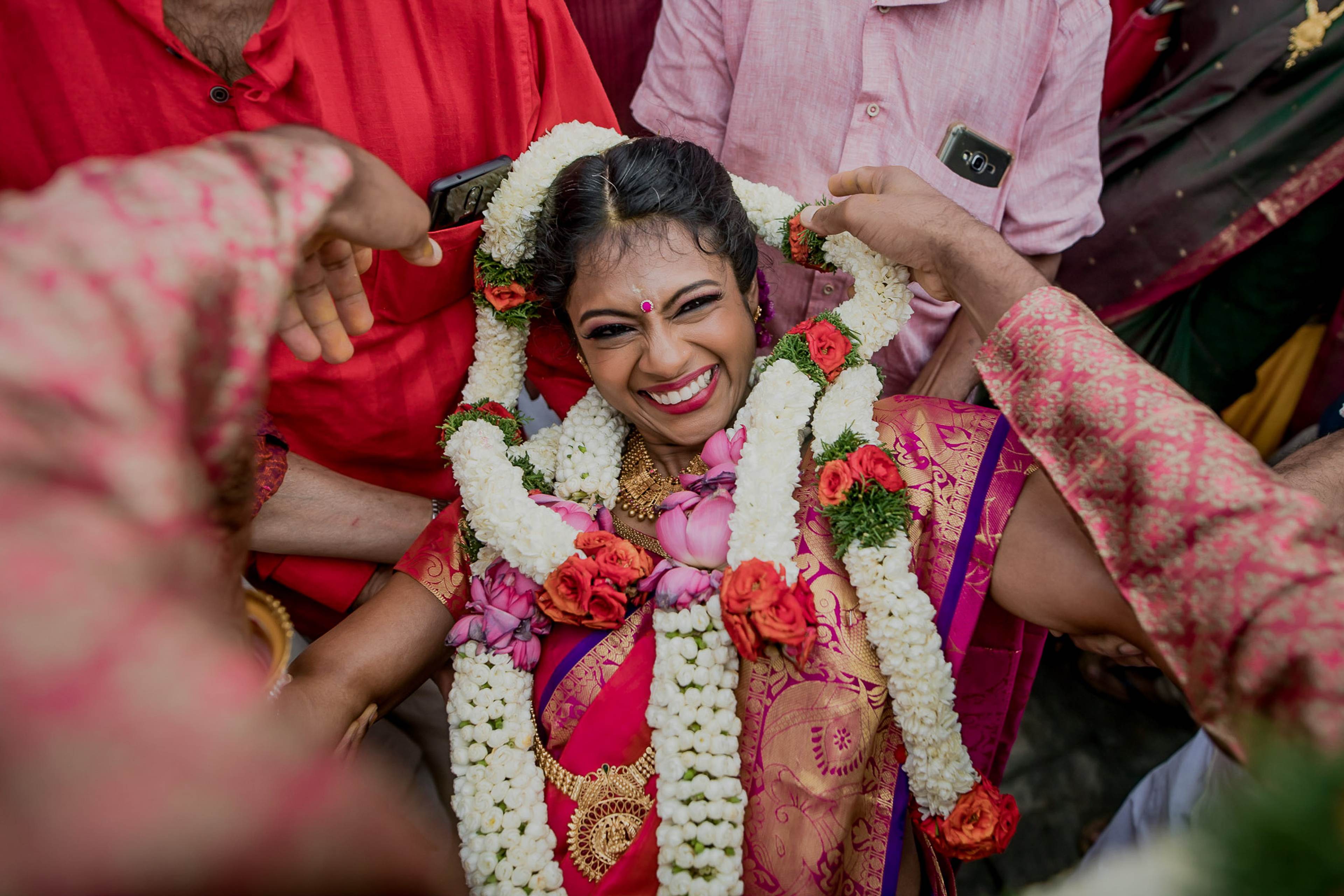 Woman in pink silk saree adorned with white and red flower garlands, surrounded by smiling family members in colorful attire