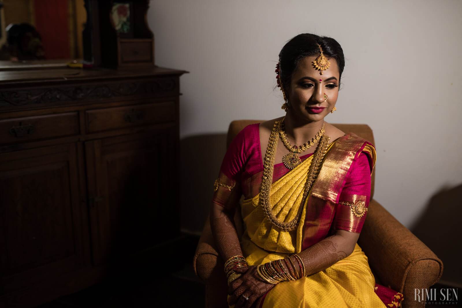 Nivetha seated in traditional South Indian bridal attire with gold jewelry and bindi, warm indoor lighting