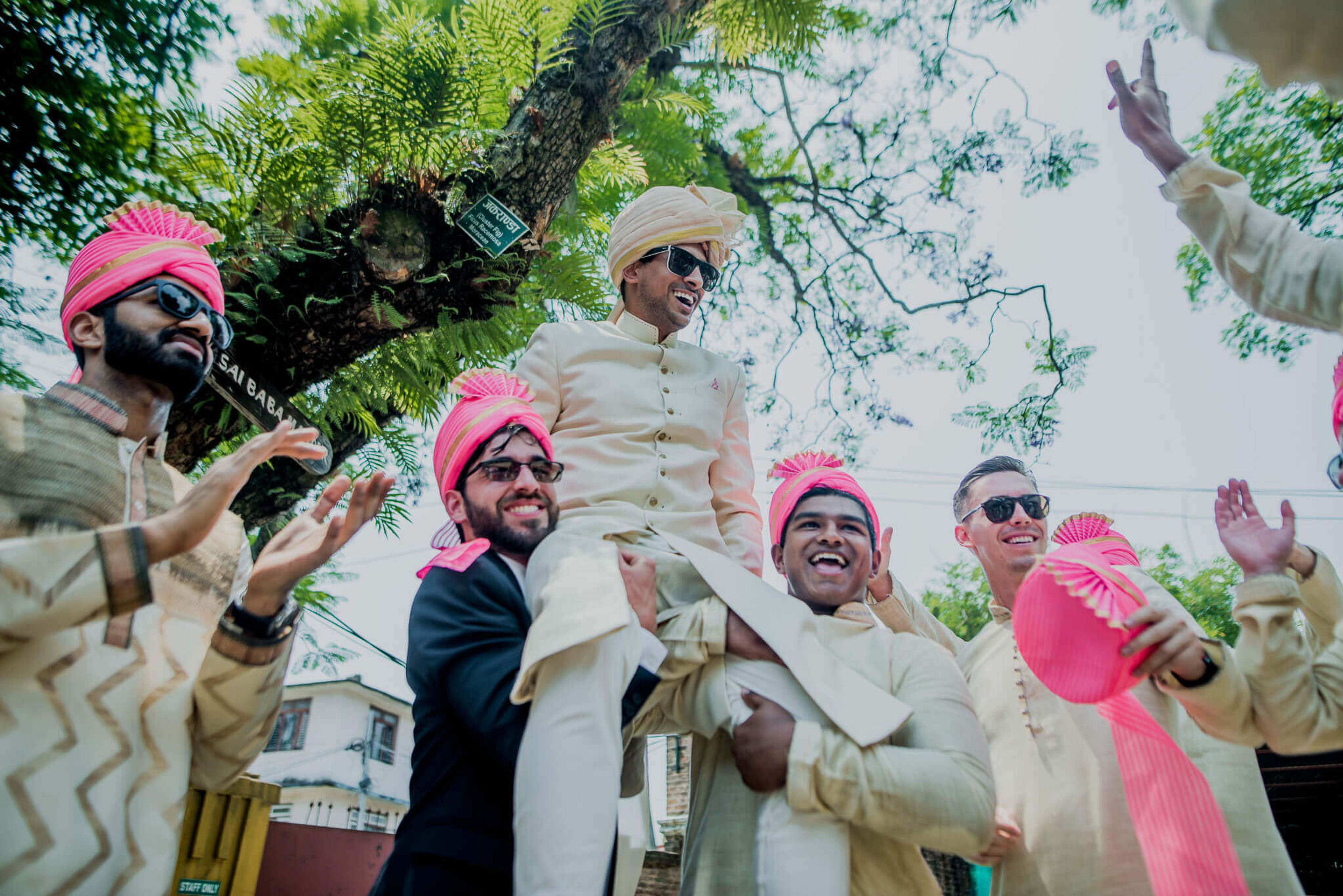Anu & Raghu celebrate with groomsmen in cream and pink attire beneath green trees in Nepal