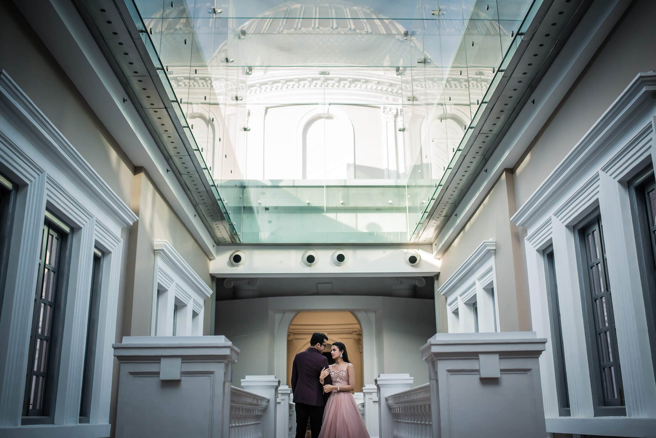 Yash and Disha stand beneath a glass skylight in Singapore's symmetrical white corridor, soft light streaming overhead