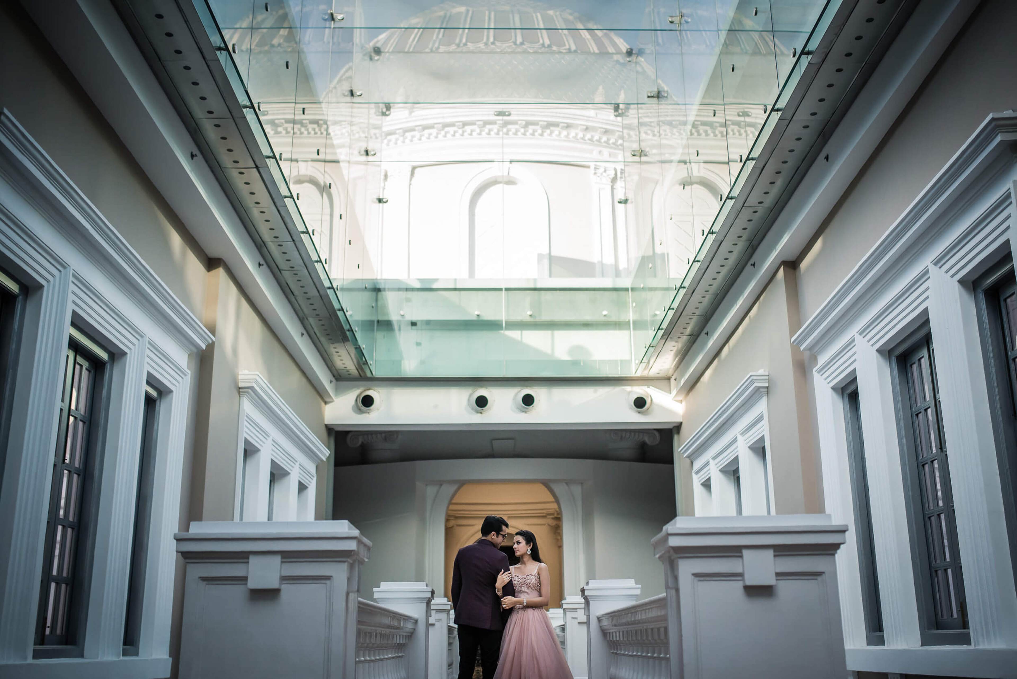 Yash and Disha stand beneath a glass skylight in Singapore's symmetrical white corridor, soft light streaming overhead