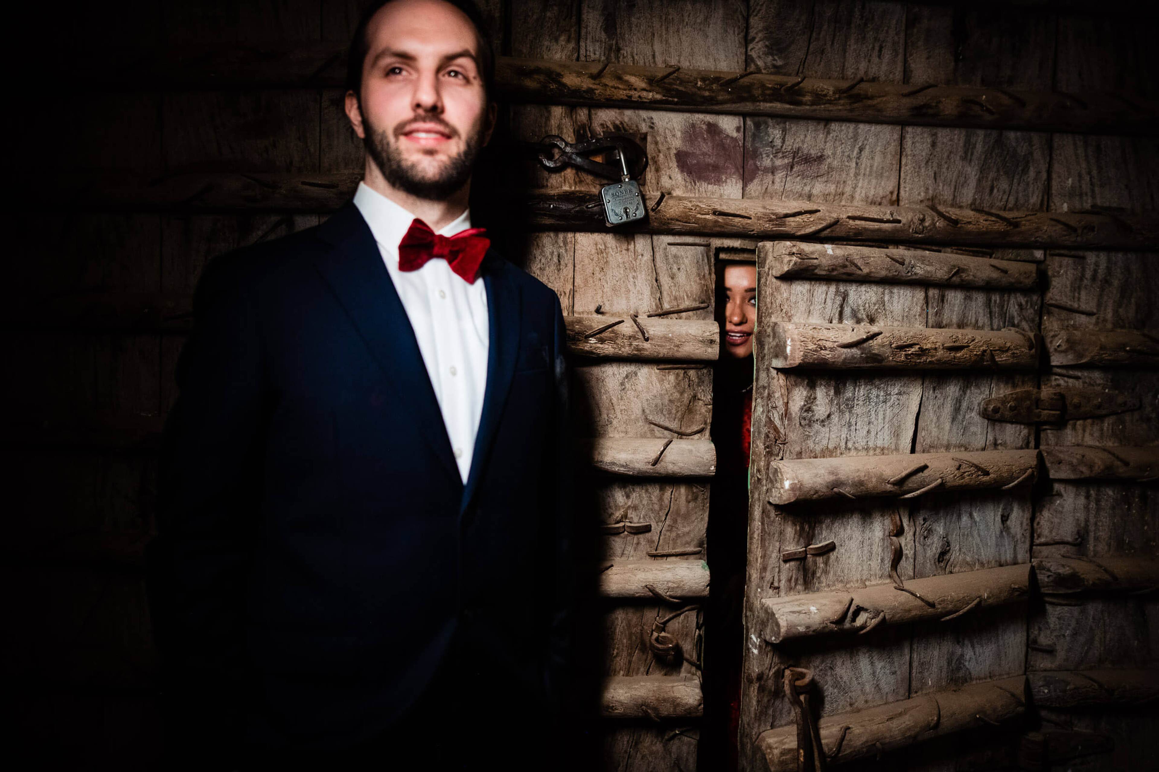 Groom in navy suit and burgundy bow tie against rustic wooden door, Rajasthan