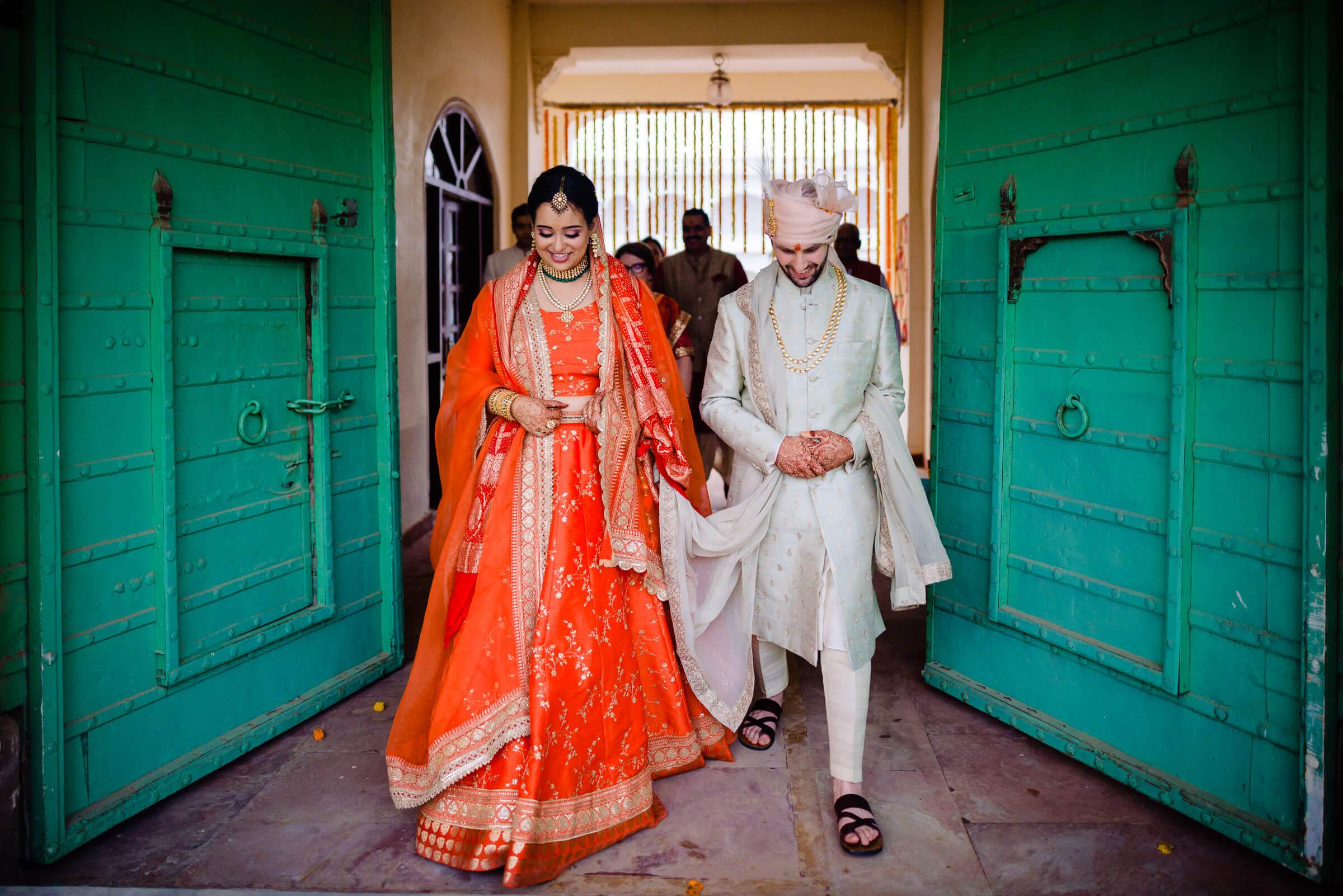 Maitrayee in red bridal lehenga and Eric in cream sherwani walk together between turquoise doors in Rajasthan