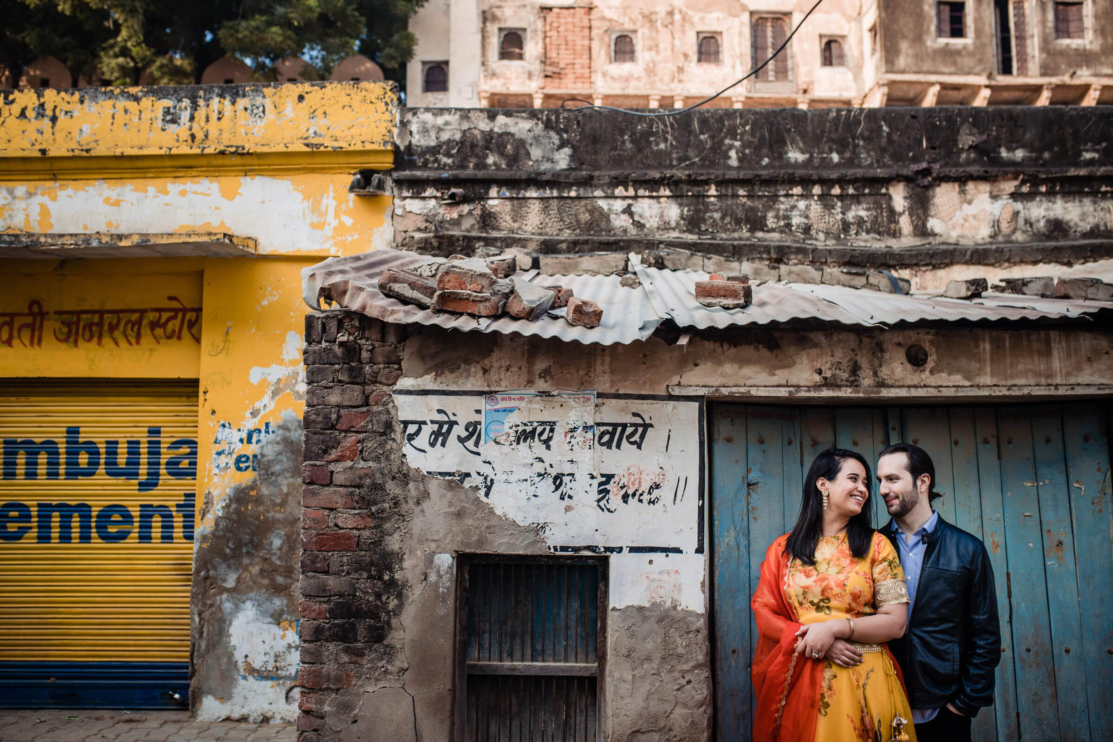 Maitrayee and Eric stand embracing against weathered Rajasthan buildings with peeling yellow and white walls