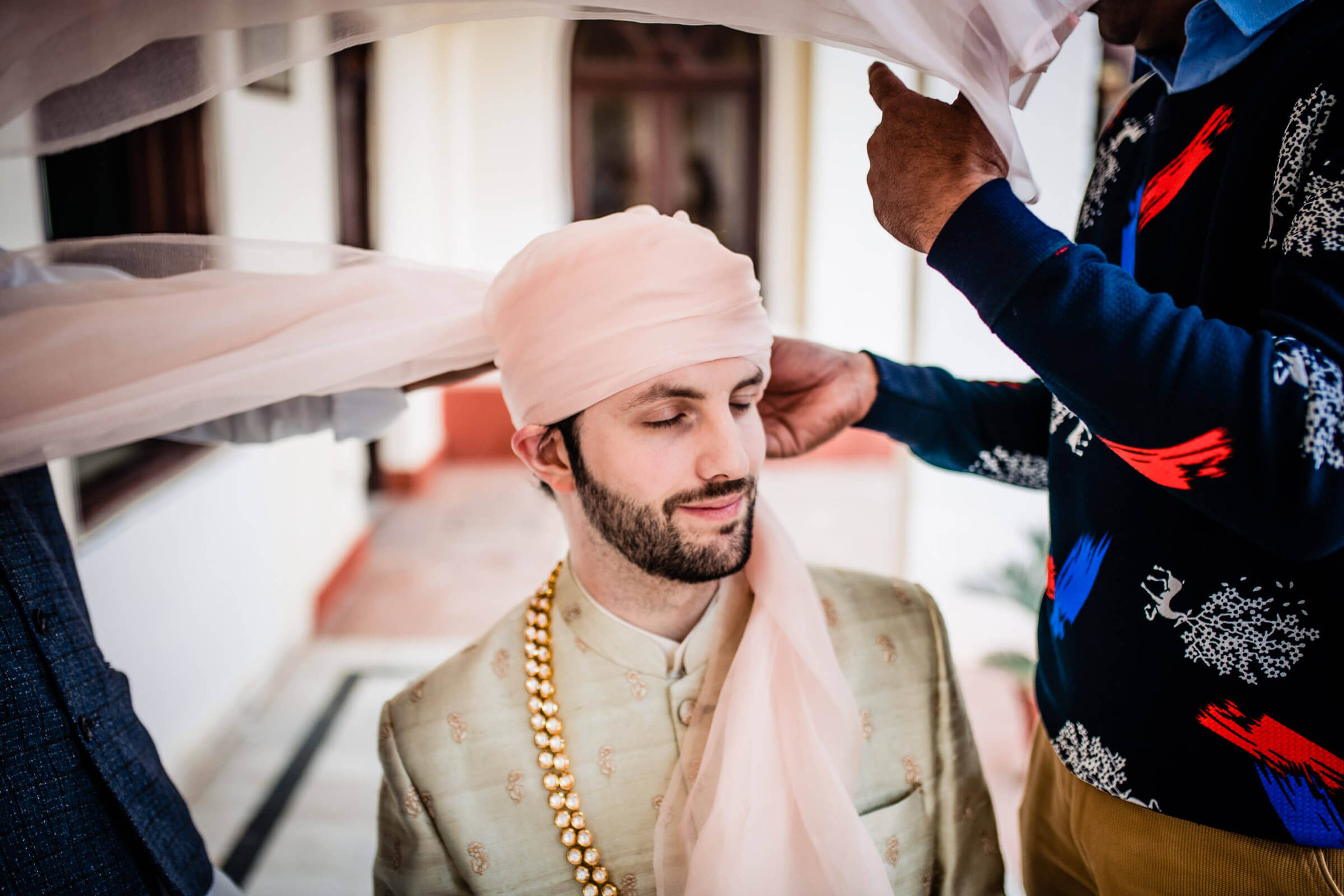Groom Eric in cream sherwani and pink turban being dressed by family in Rajasthan