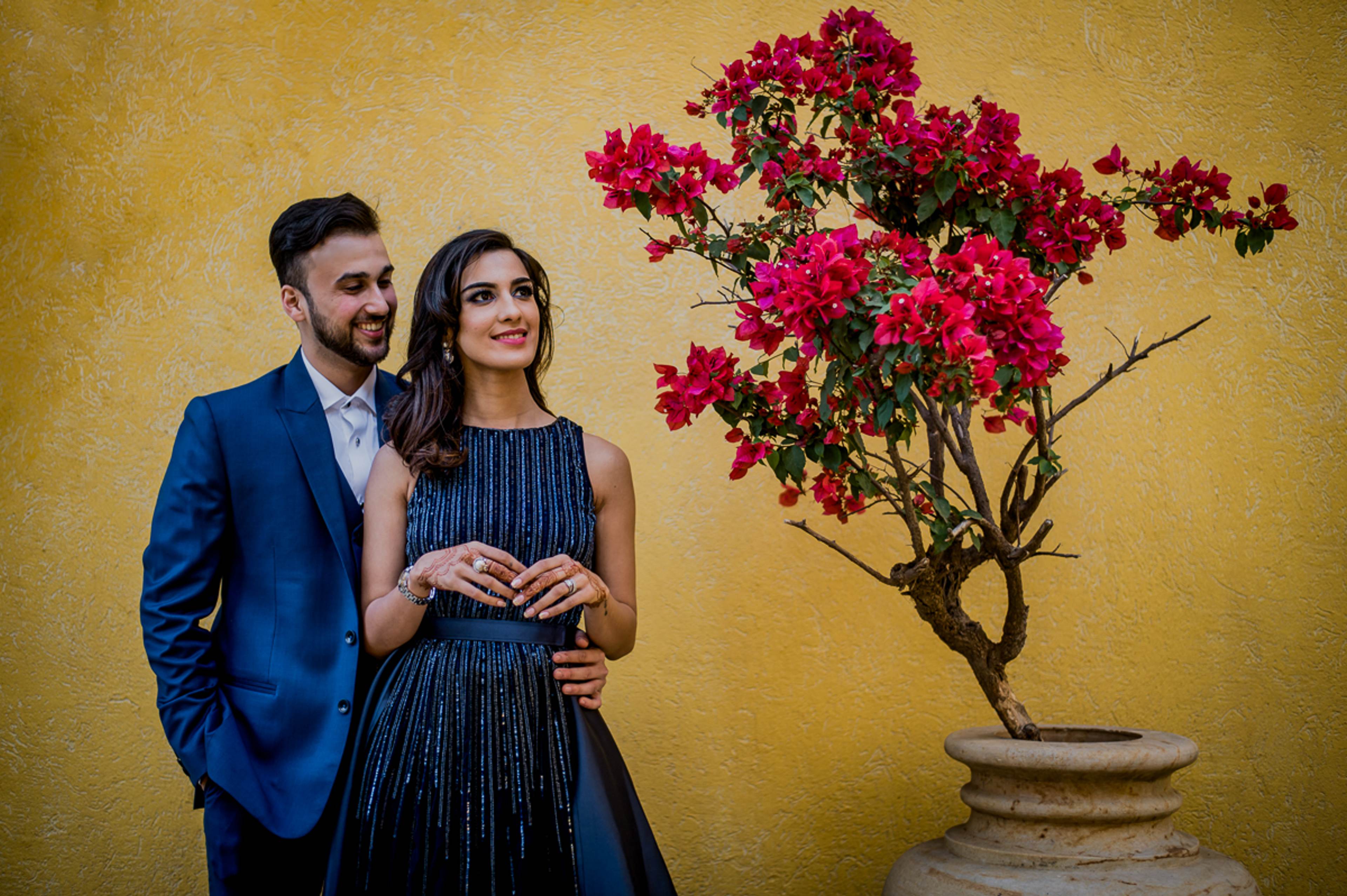 Couple in blue blazer and navy dress pose beside blooming magenta bougainvillea against golden yellow wall