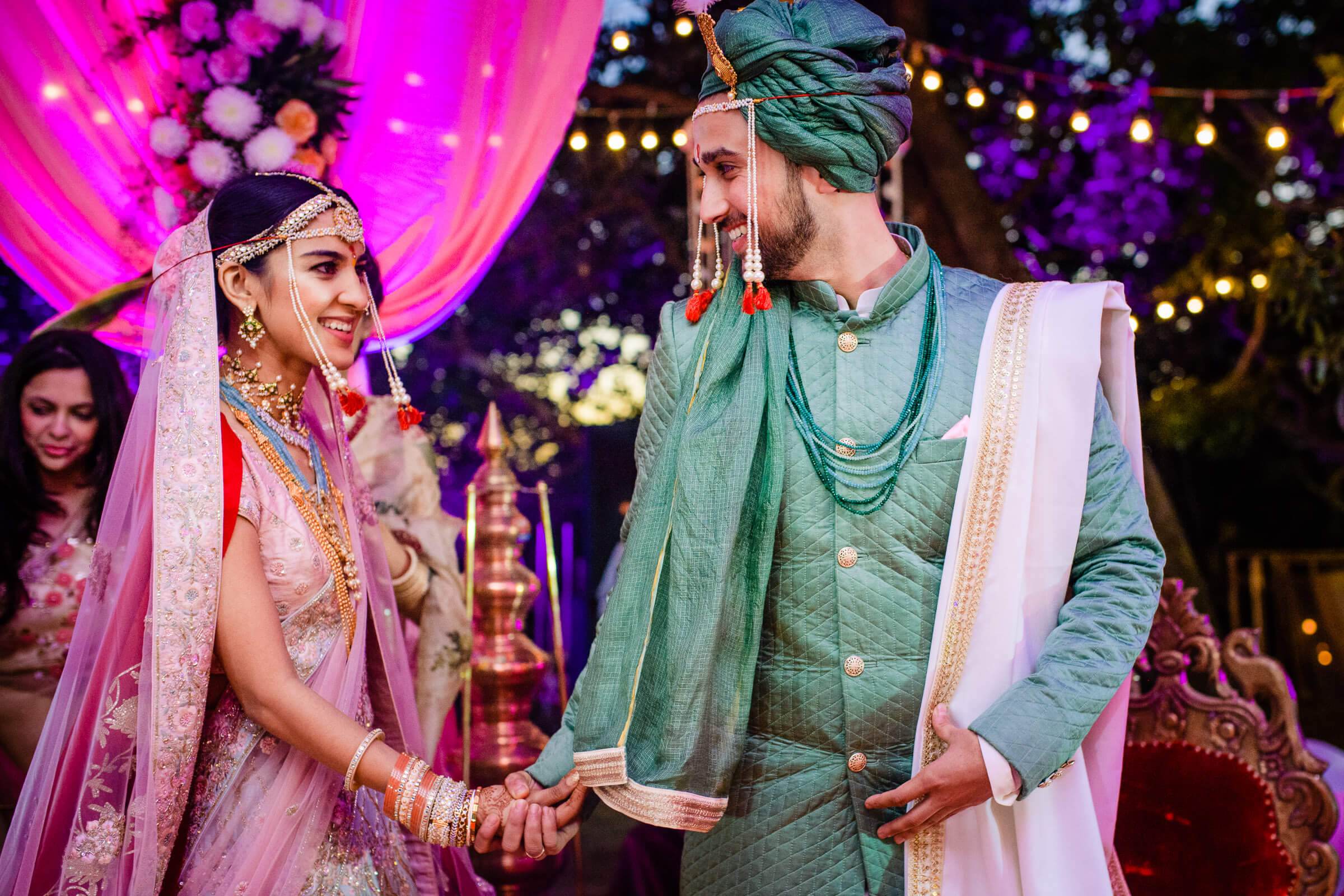 Shreya and Yash hold hands during their wedding ceremony in traditional attire under vibrant pink lighting and string lights at Mahabaleshwar