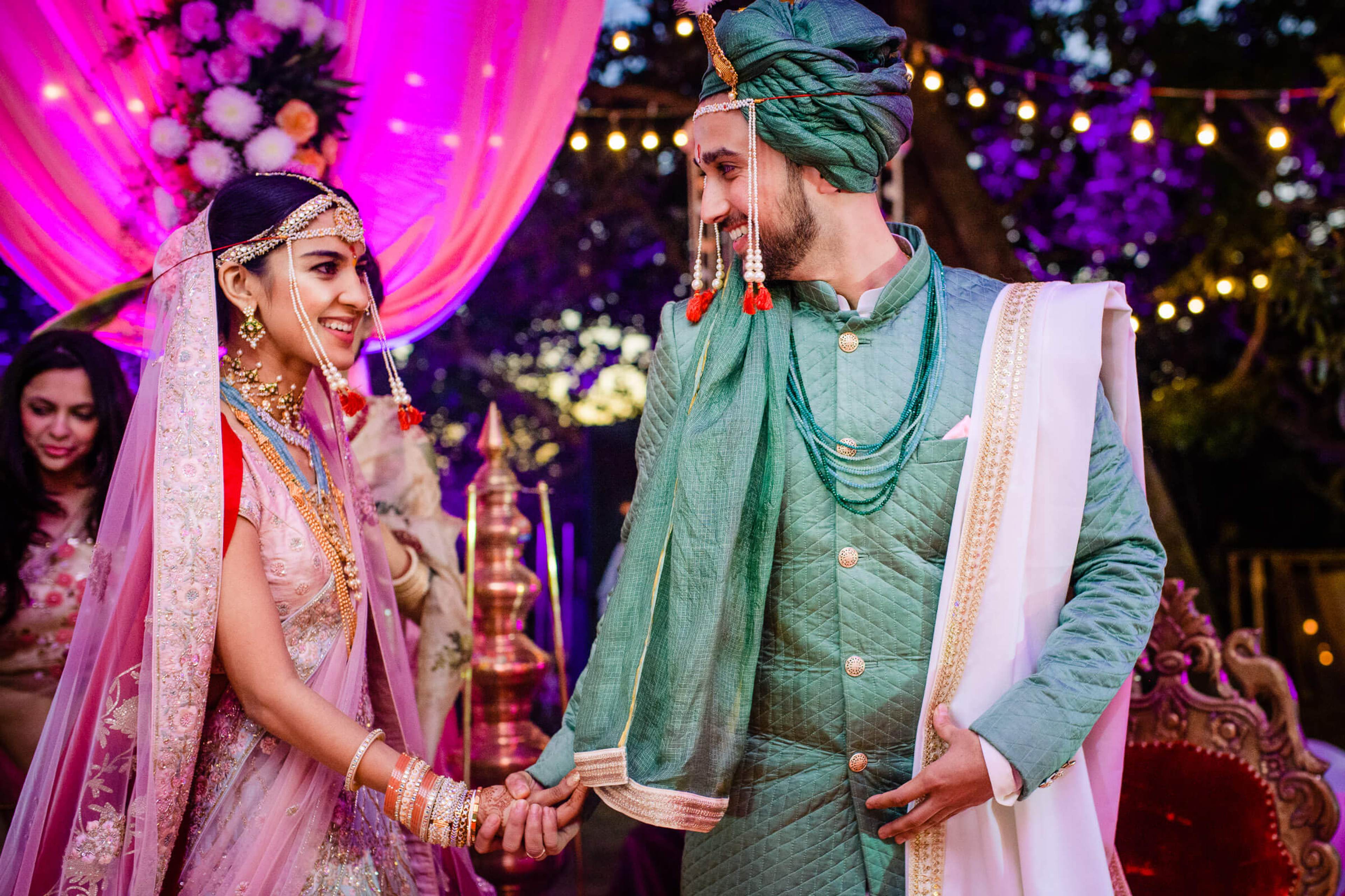 Shreya and Yash hold hands during their wedding ceremony in traditional attire under vibrant pink lighting and string lights at Mahabaleshwar