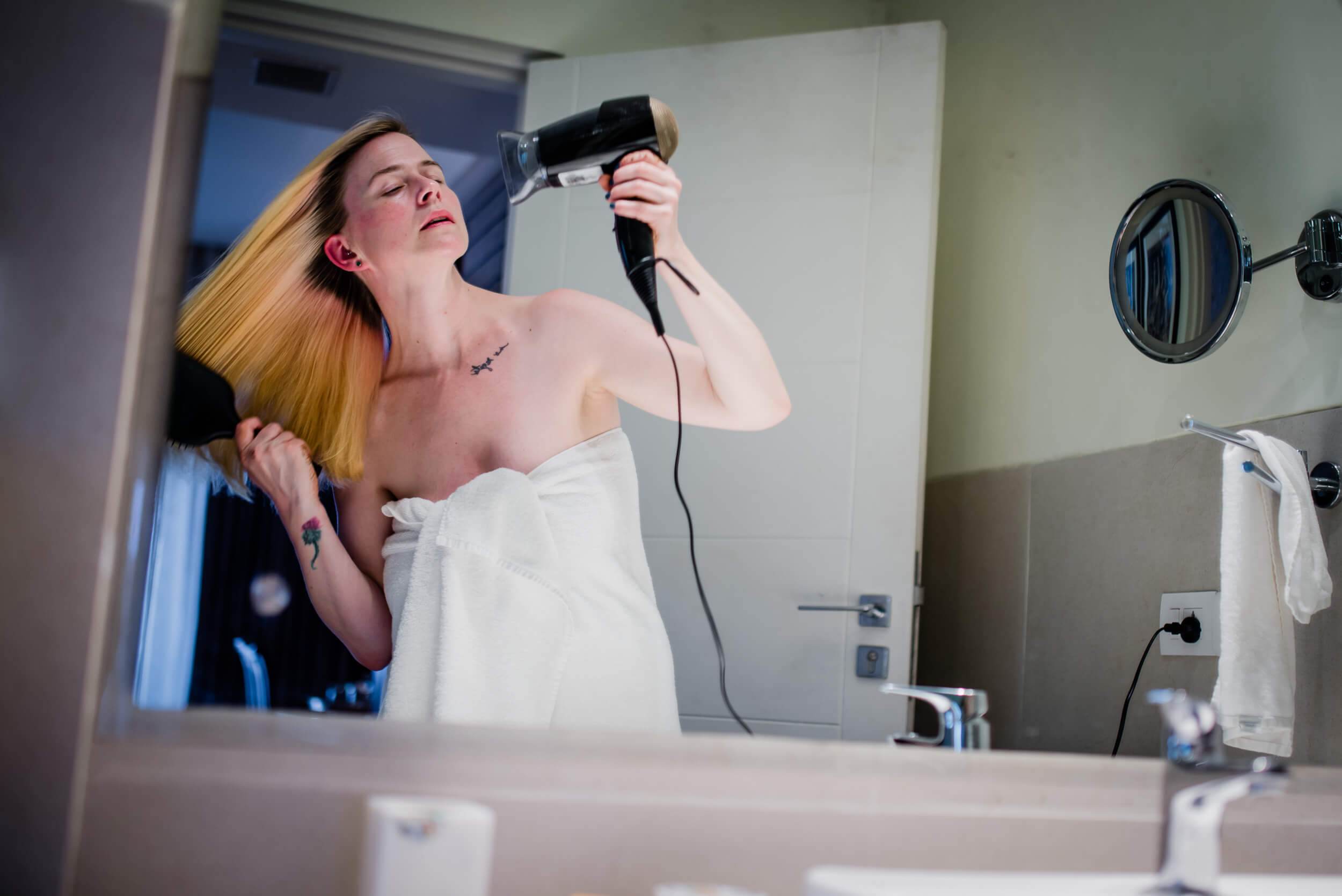 Sarah drying her hair with a blow dryer in a bathroom mirror, wrapped in white towel