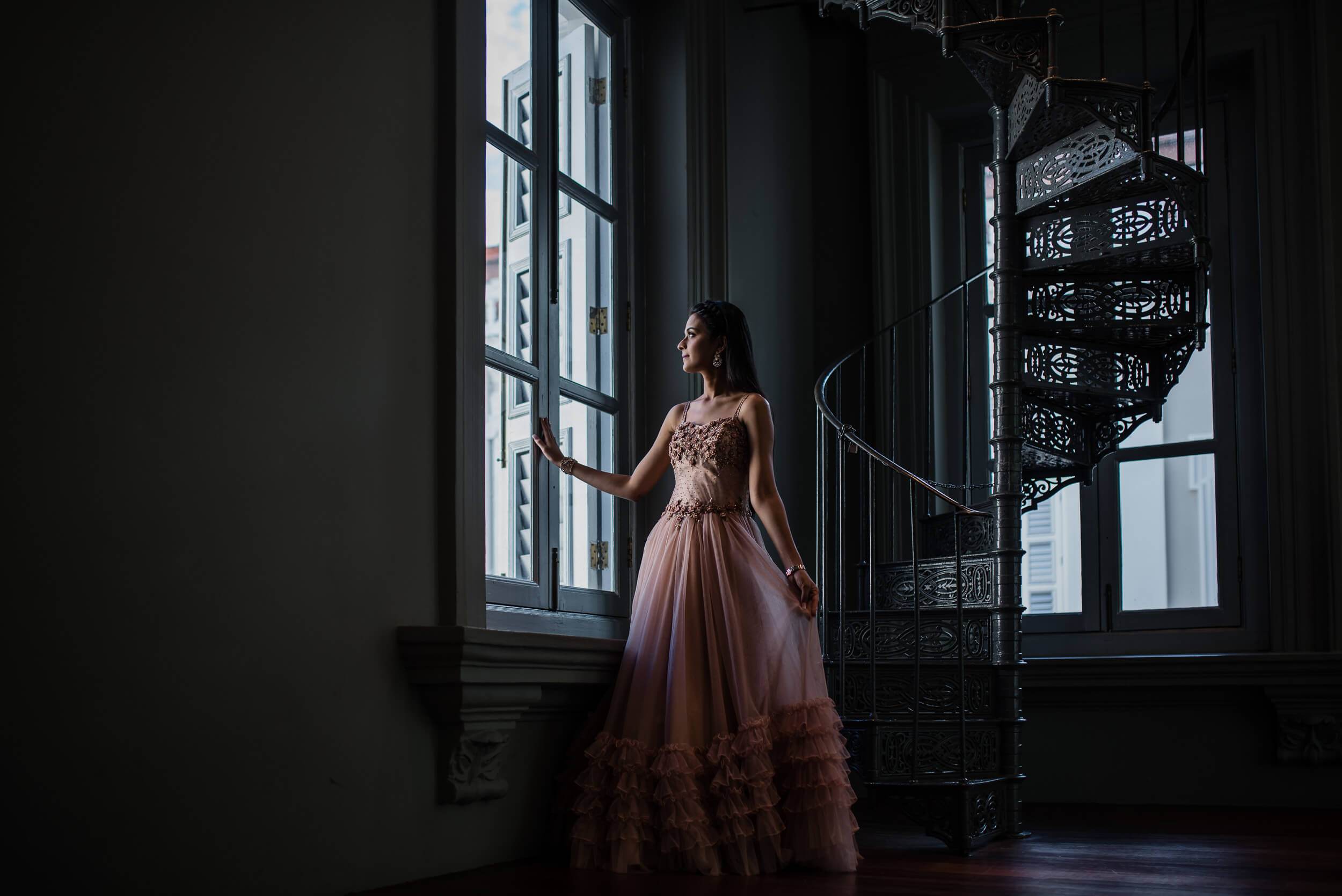 Disha in blush tulle gown gazing from industrial Singapore loft window, architectural metal spiral staircase visible