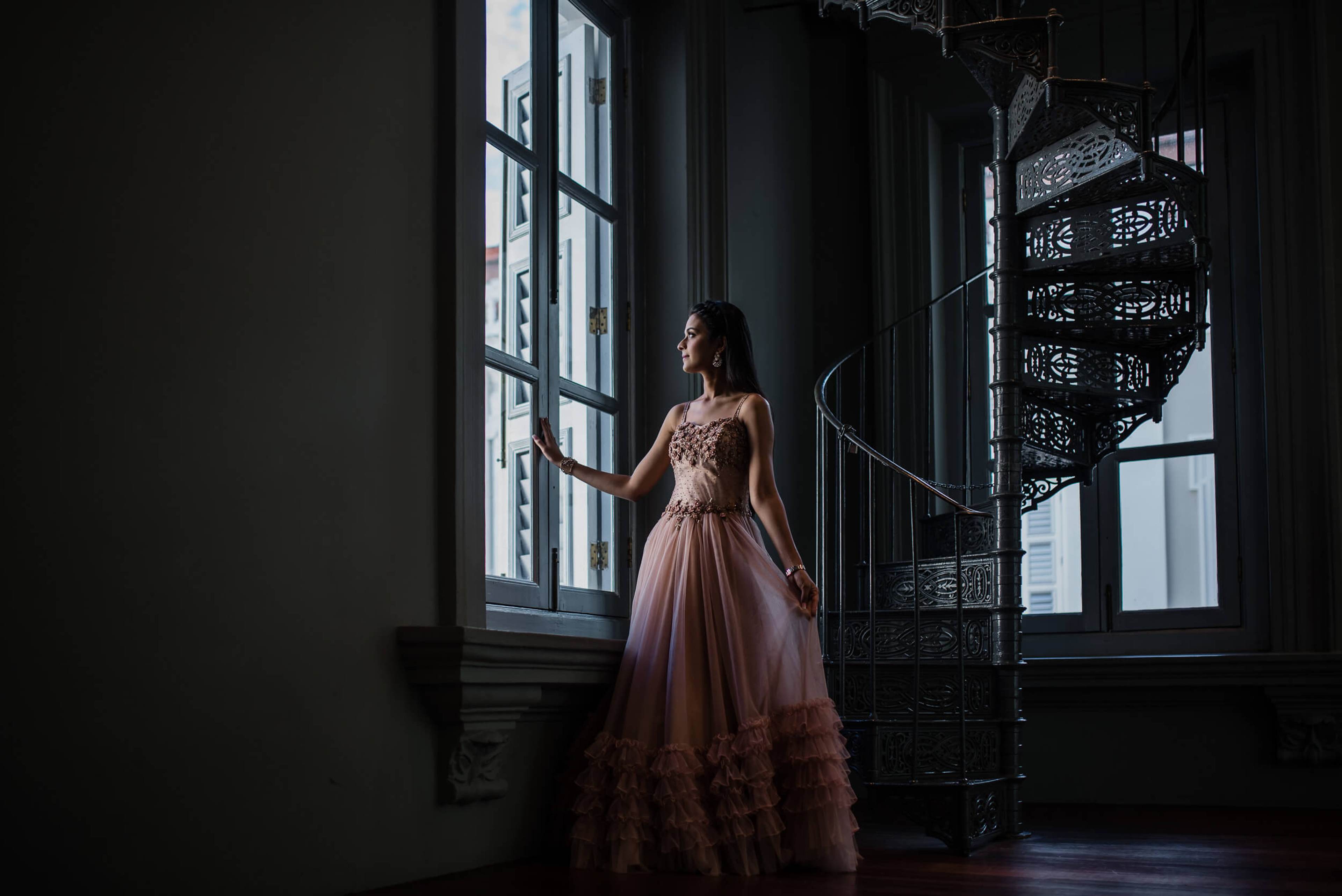 Disha in blush tulle gown gazing from industrial Singapore loft window, architectural metal spiral staircase visible