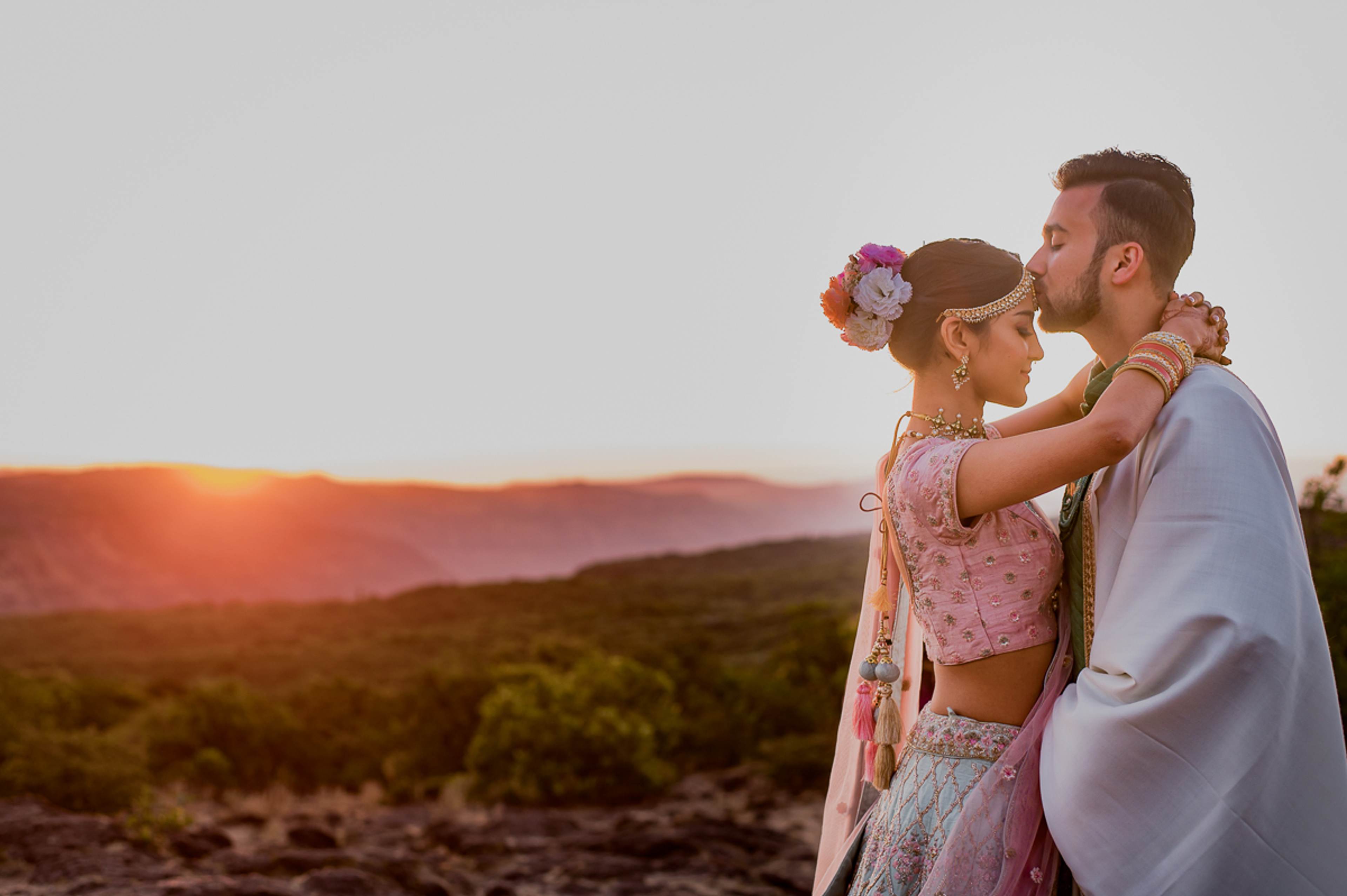 Couple in pink and lavender attire with flower crown embracing at sunset over rolling landscape