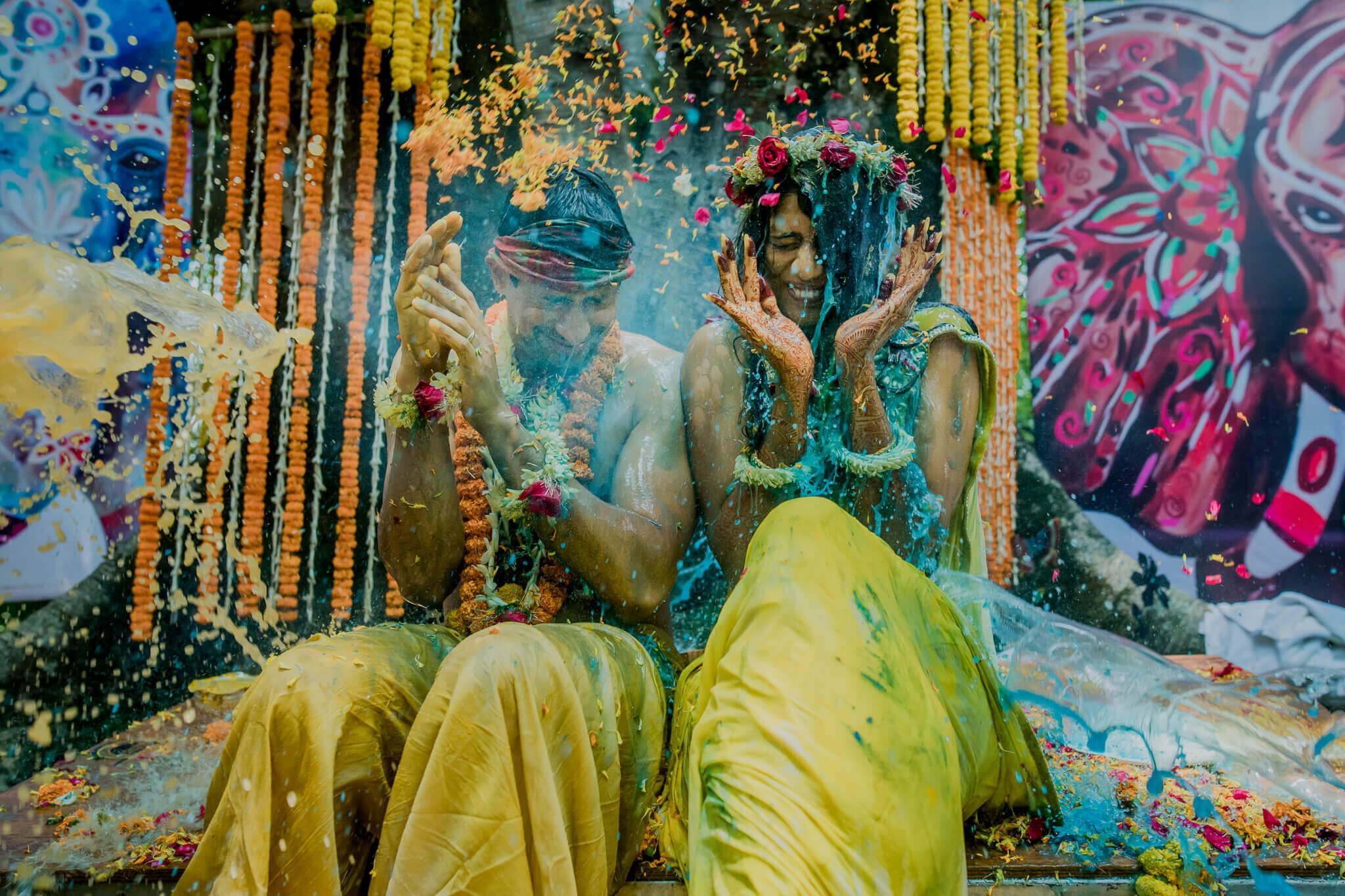Anu and Raghu covered in colored powder during Holi celebration with marigold garlands and vibrant backdrop in Nepal