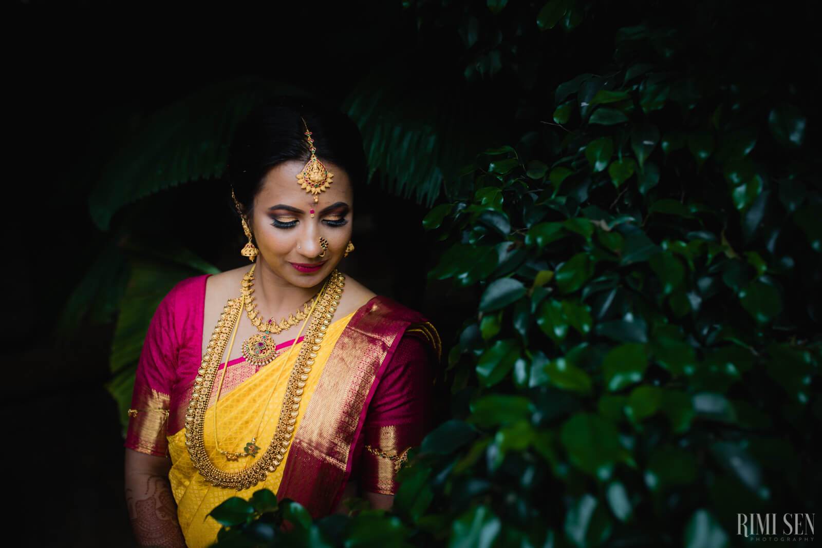Woman in traditional yellow and magenta silk saree with gold jewelry surrounded by green foliage, Bangalore