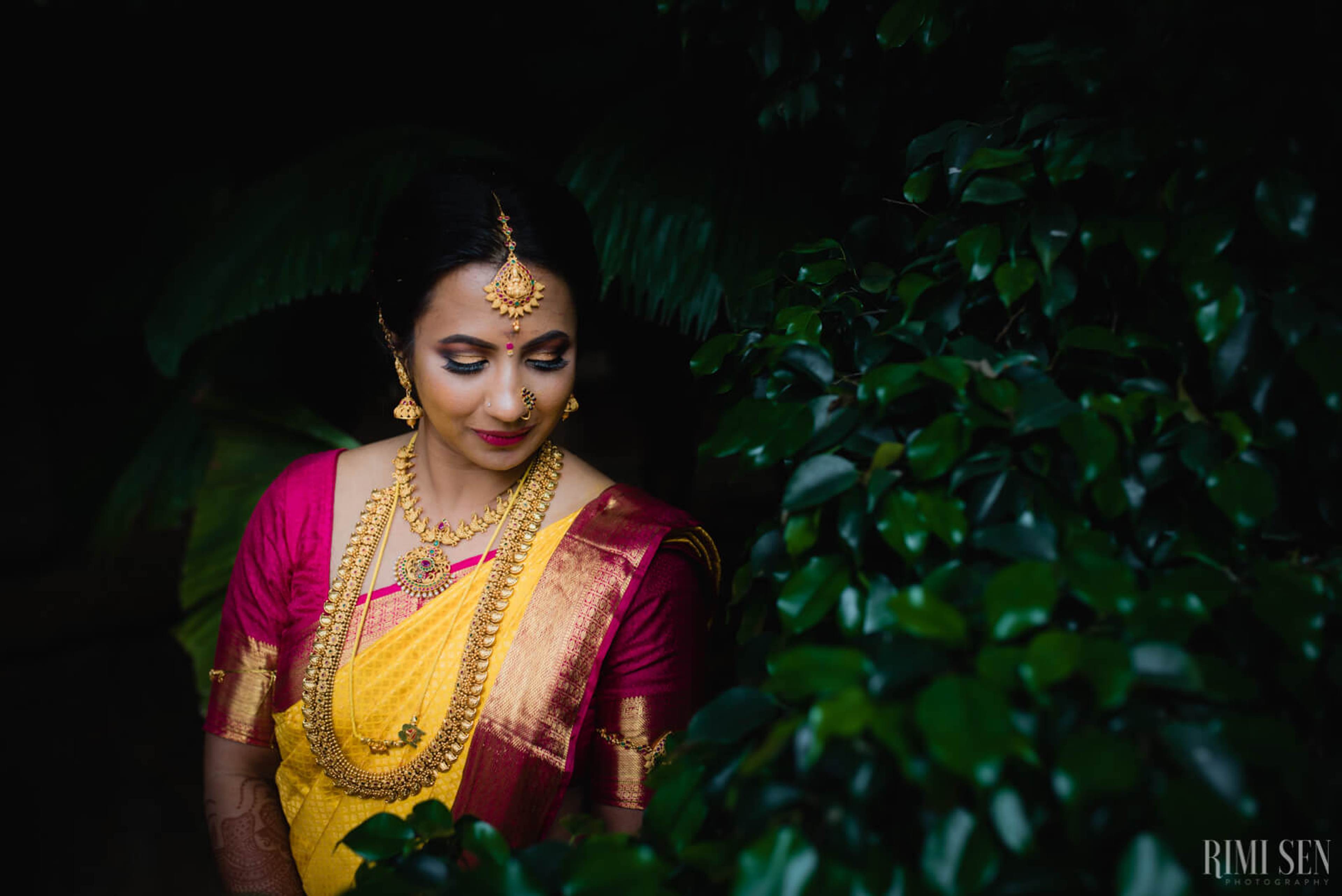 Woman in traditional yellow and magenta silk saree with gold jewelry surrounded by green foliage, Bangalore