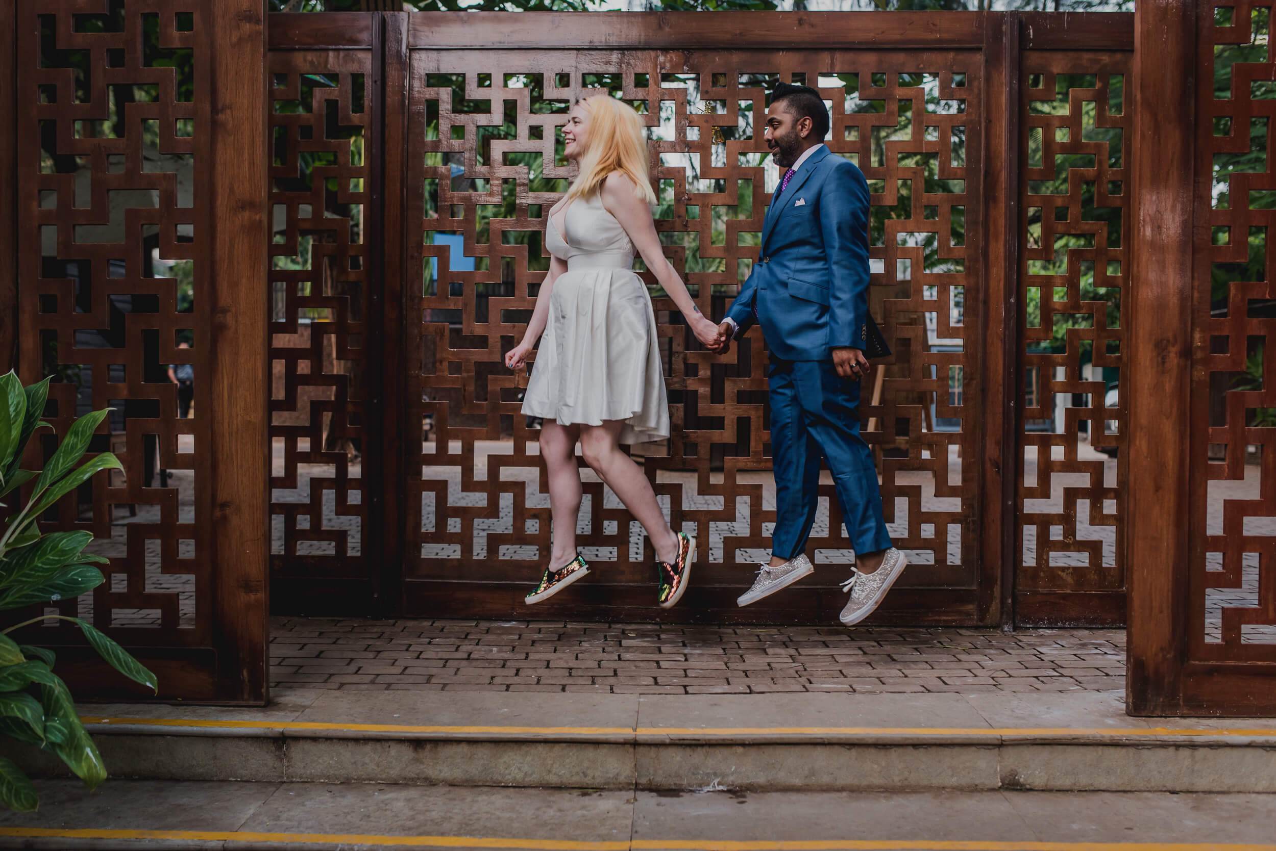 Sarah and Ravin jump hand-in-hand before ornate wooden panels in Goa, both mid-air, smiling