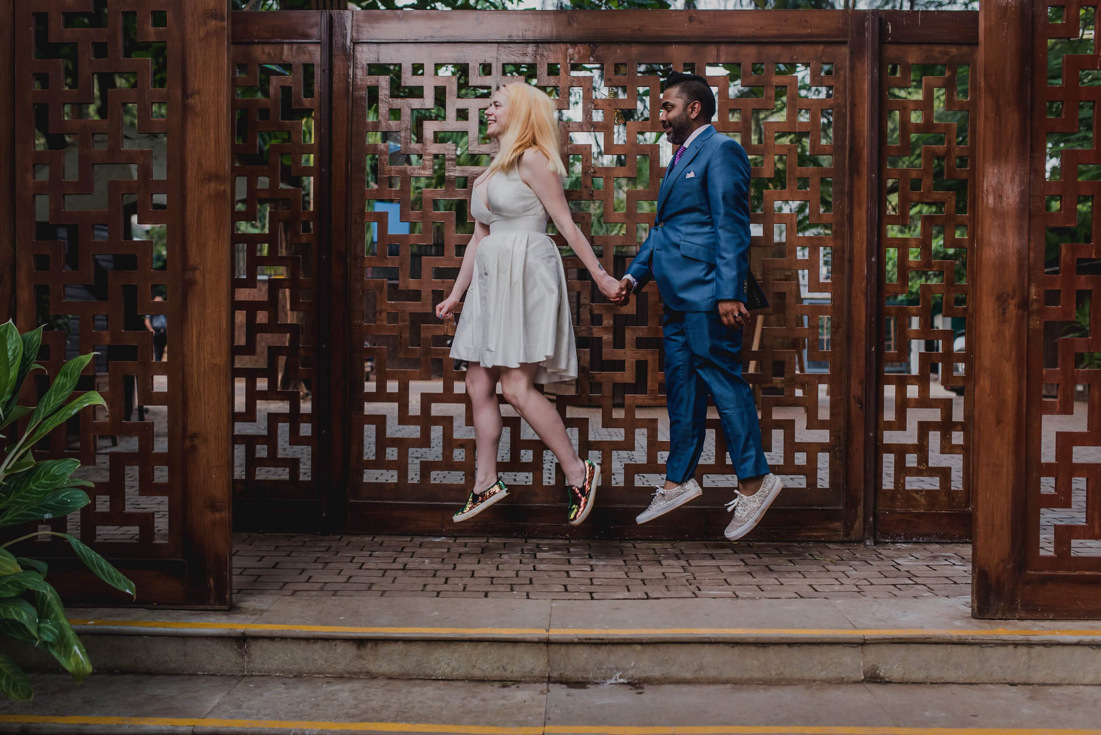 Sarah and Ravin jump hand-in-hand before ornate wooden panels in Goa, both mid-air, smiling
