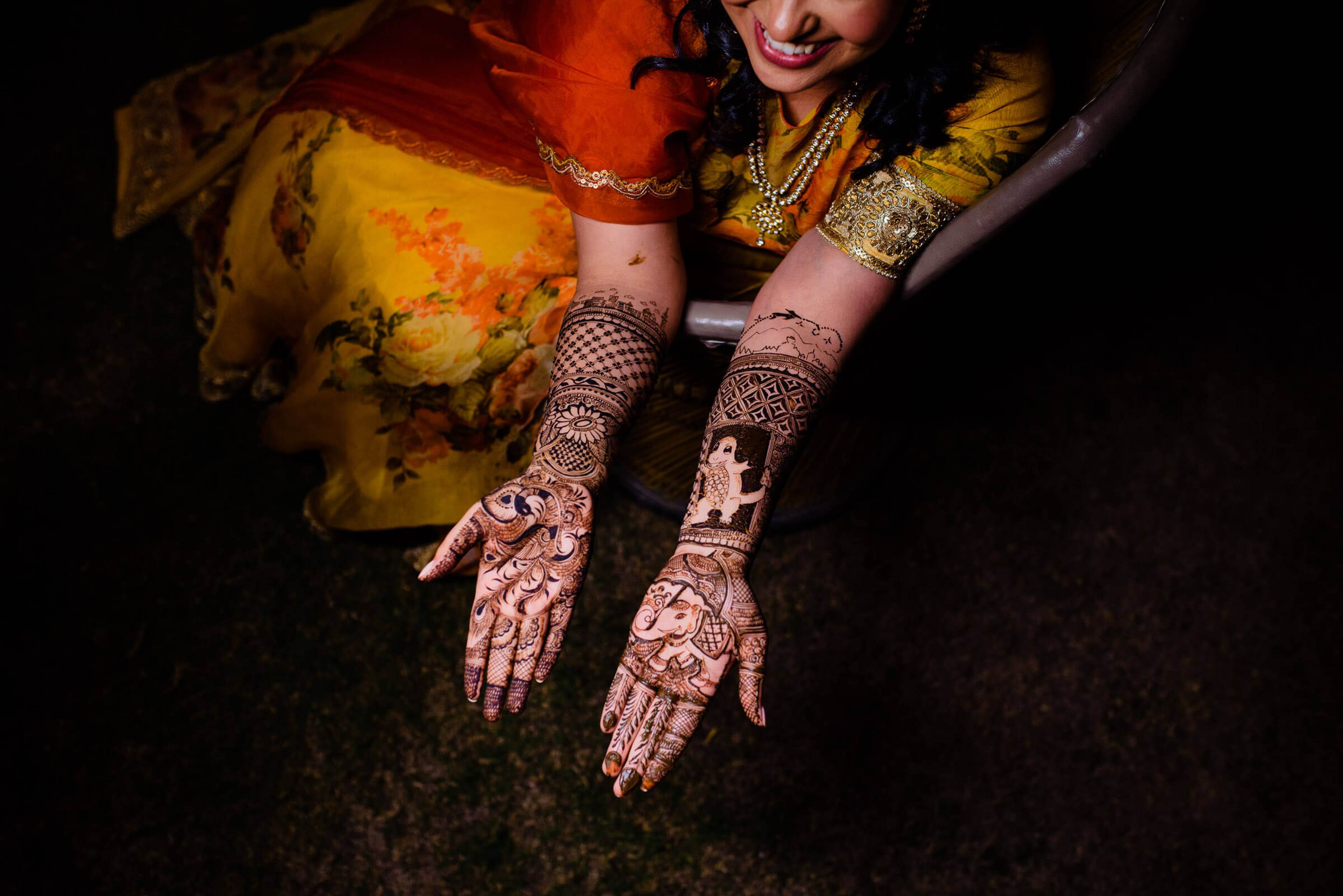 Maitrayee displays intricate henna on hands and arms, wearing gold jewelry and yellow silk against dark background