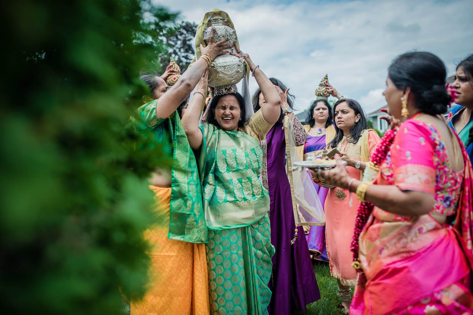 Barkha lifts a decorated pot above her head while surrounded by family in colorful traditional attire at New York celebration