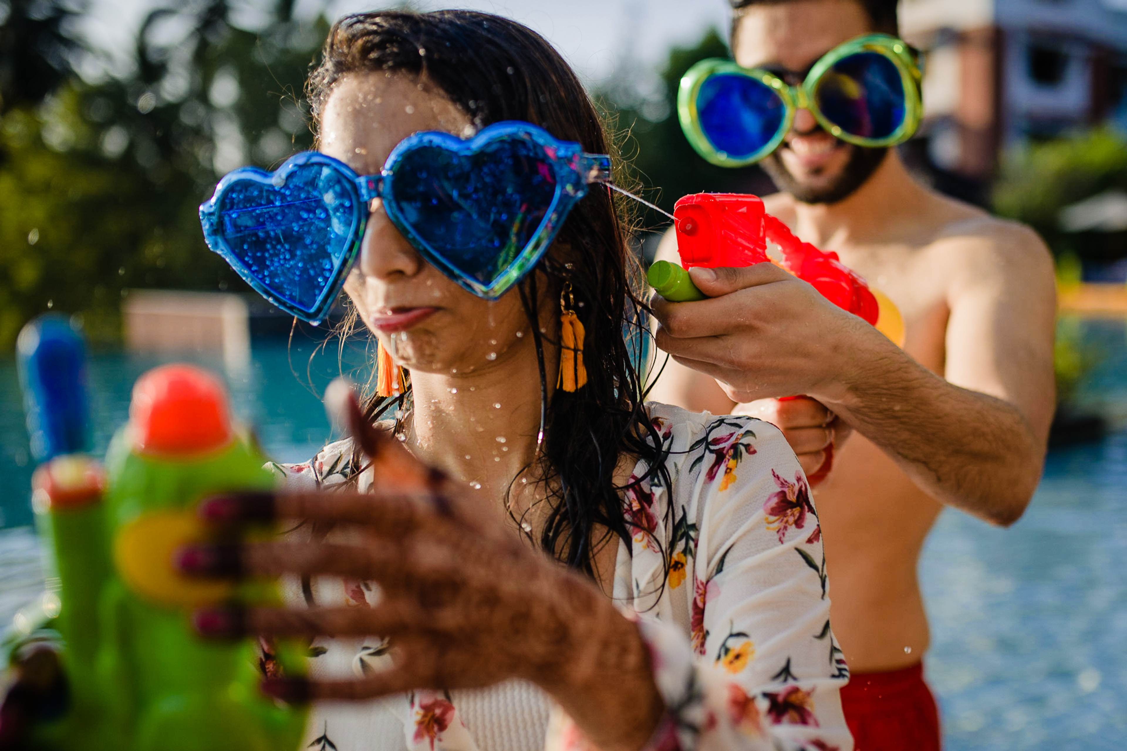 Avani & Srikant laugh in a swimming pool, wearing novelty heart-shaped sunglasses and playing with water guns