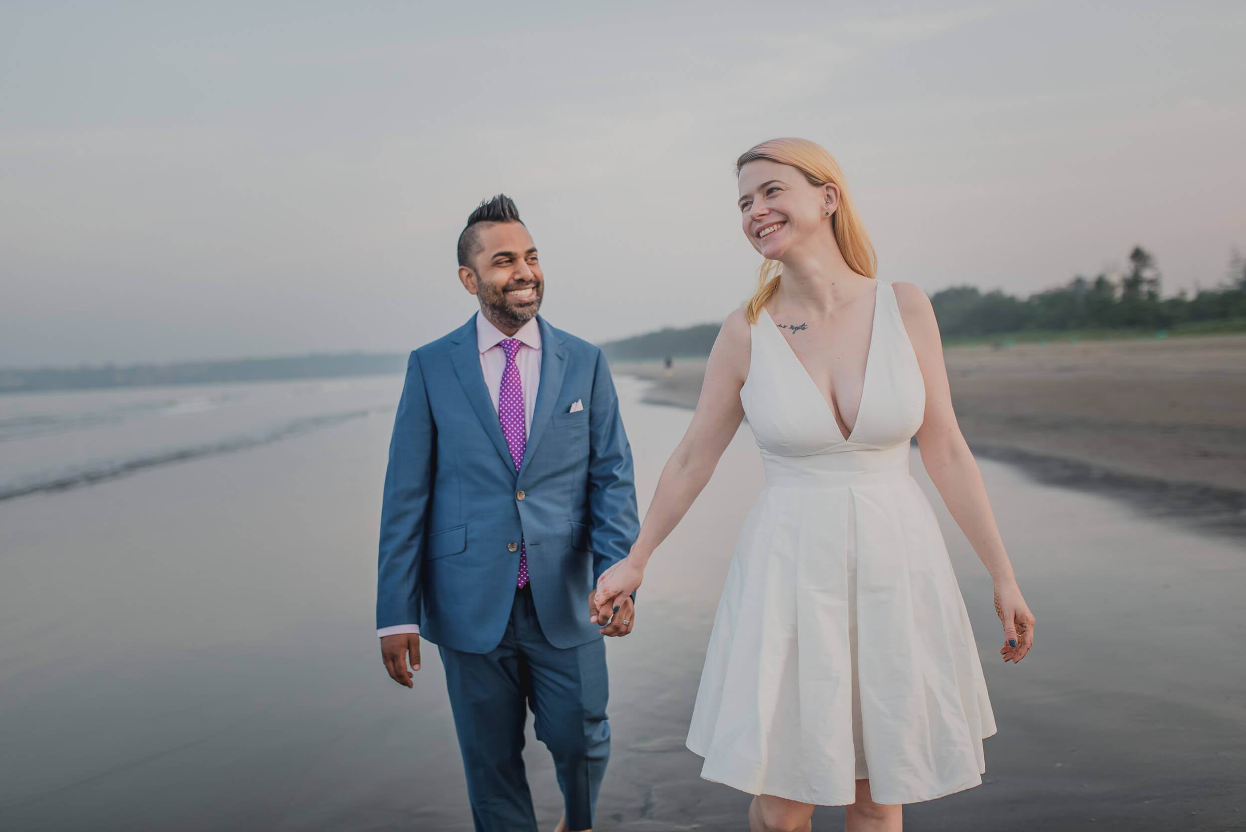 Sarah and Ravin hold hands walking along Goa's beach at dusk, smiling toward horizon