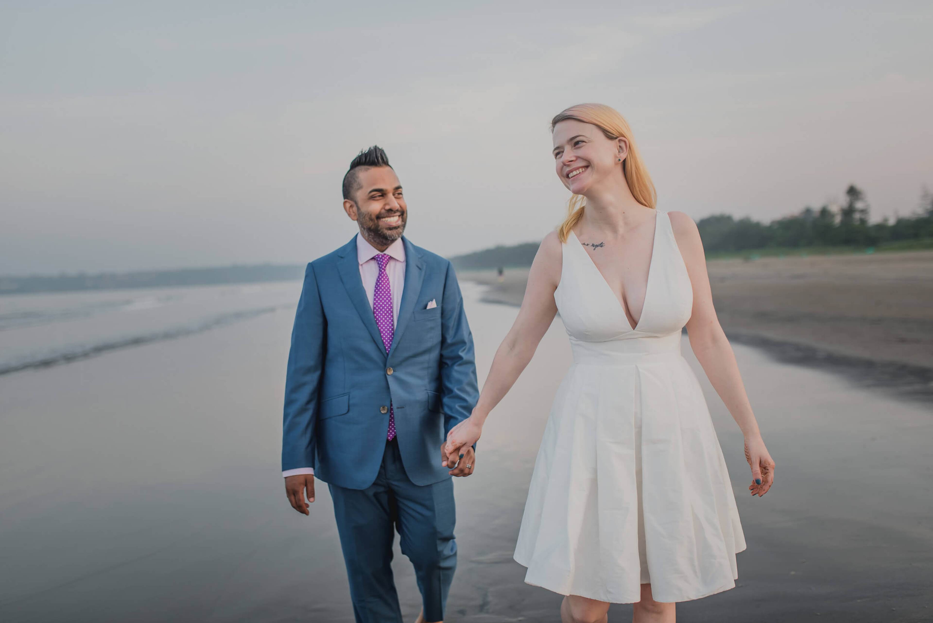 Sarah and Ravin hold hands walking along Goa's beach at dusk, smiling toward horizon