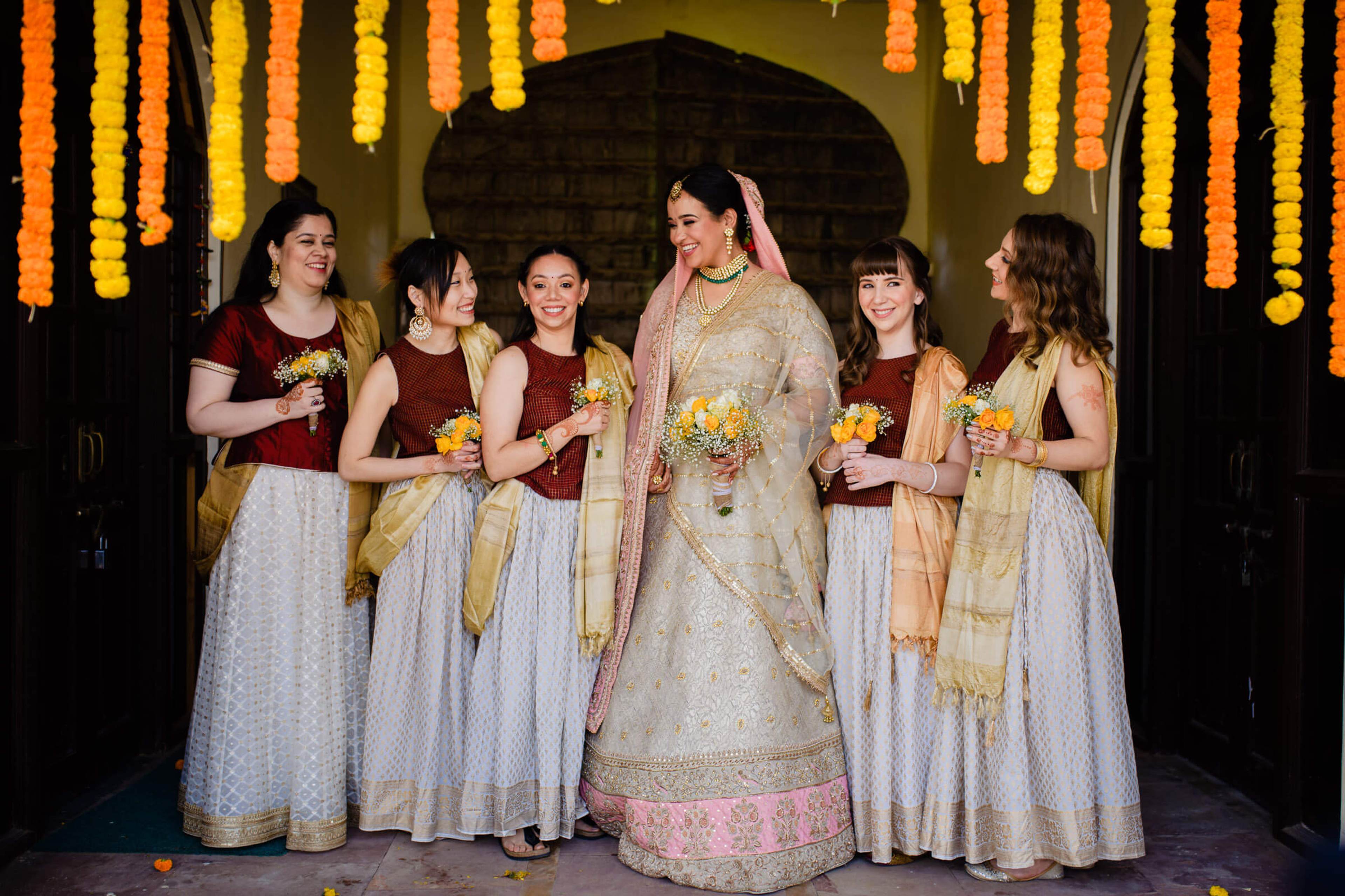 Maitrayee in embroidered lehenga with bridesmaids in maroon and gold, holding yellow flowers under marigold garlands in Rajasthan