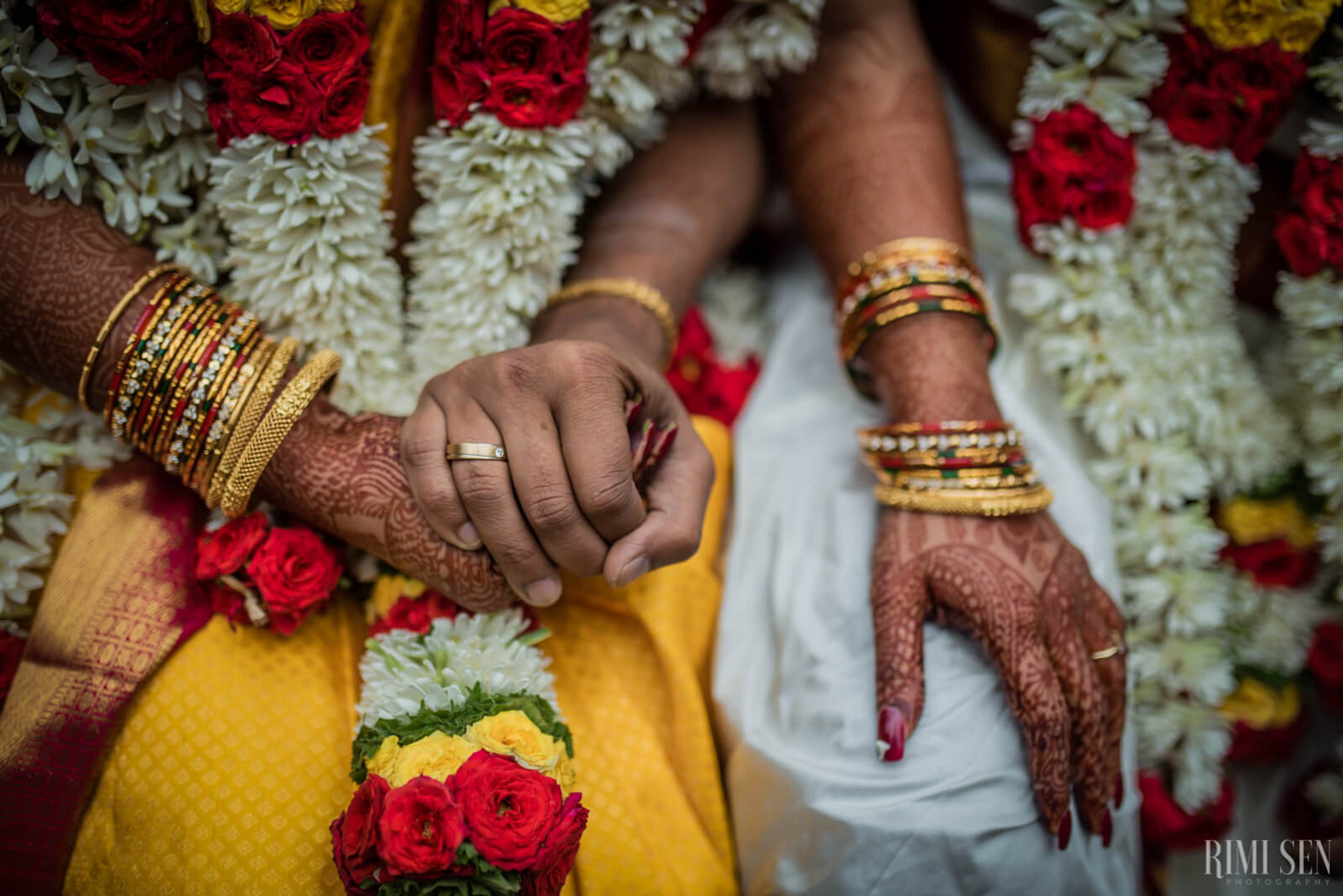 Nivetha & Sridhar hold hands adorned with gold bangles and henna, surrounded by red and white flowers in Bangalore