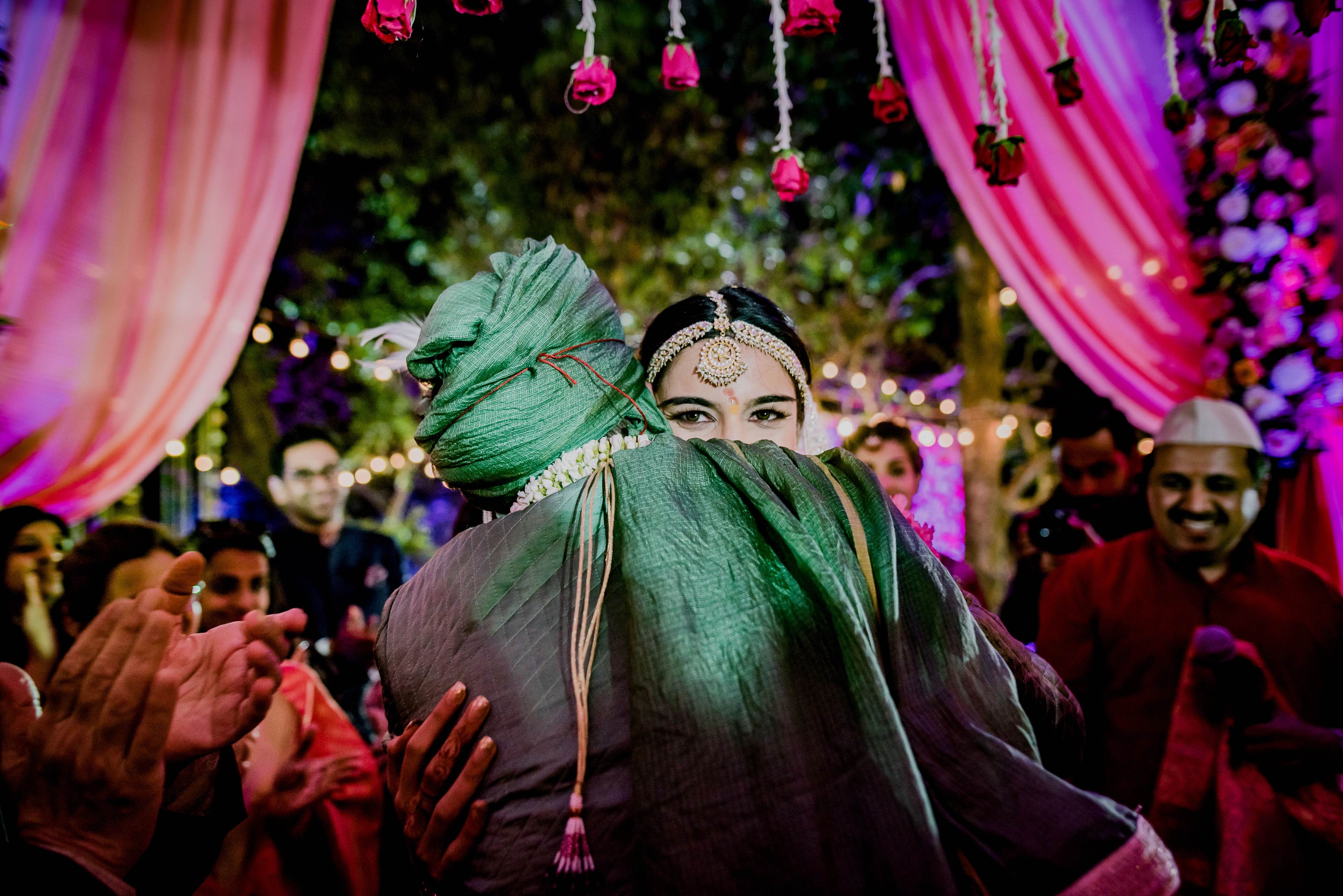 Couple in traditional attire surrounded by guests under pink fabric and hanging florals at vibrant celebration