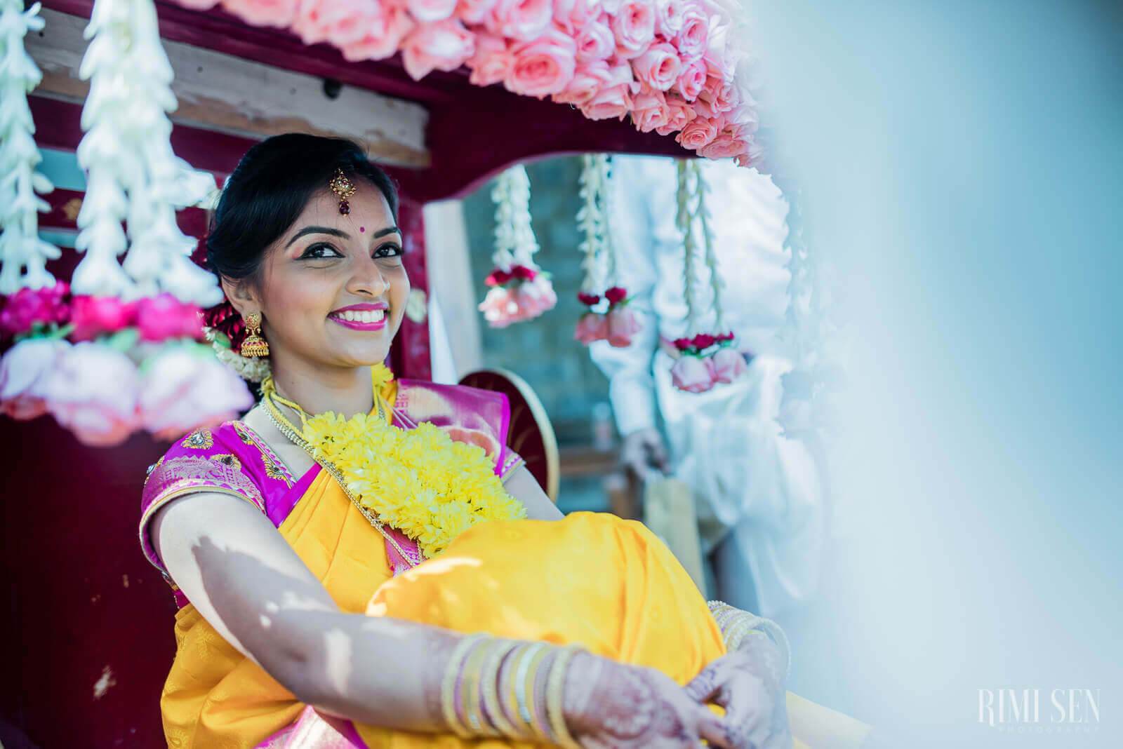 Pooja smiles beneath pink floral canopy, wearing yellow and magenta wedding attire with jewelry