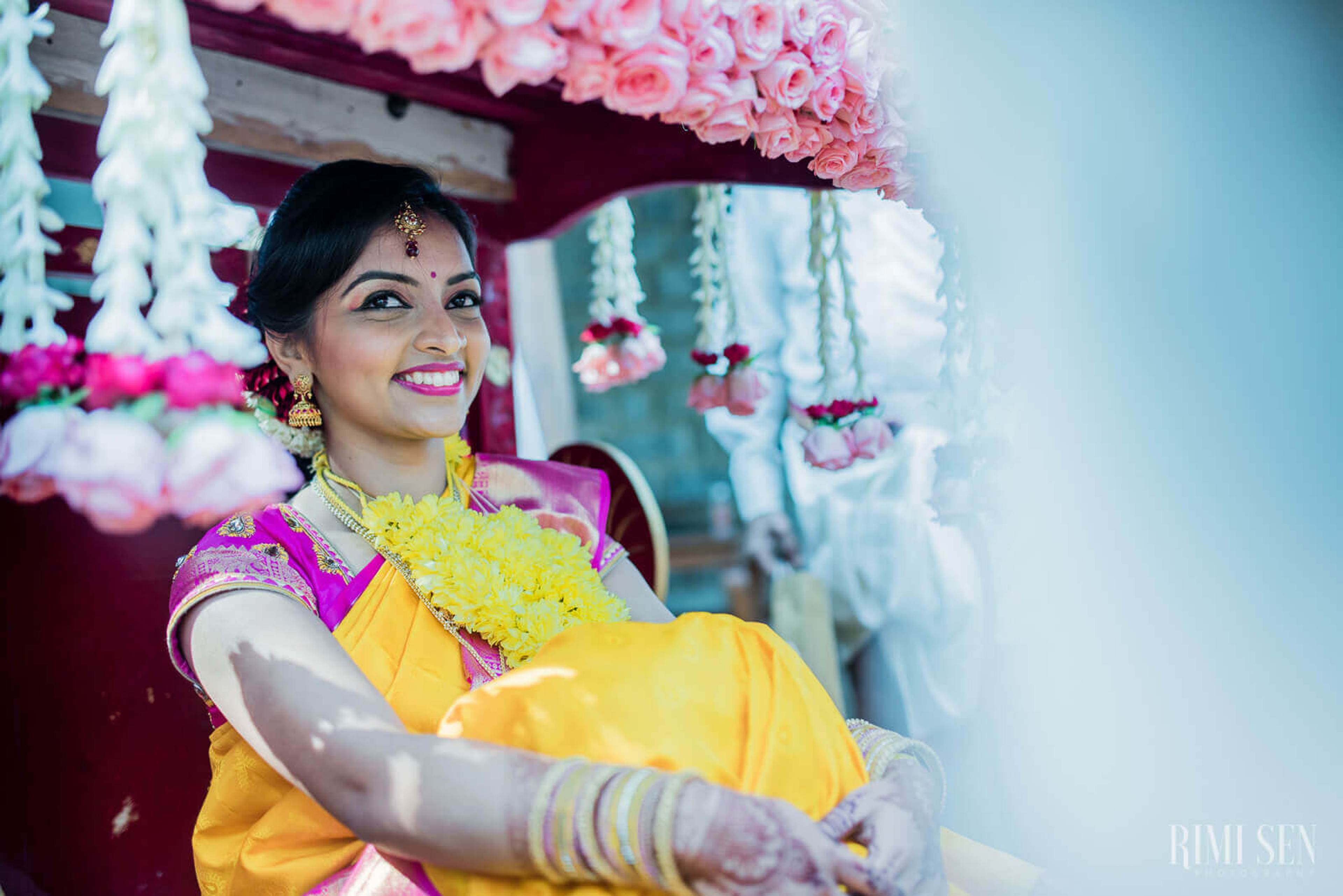 Pooja smiles beneath pink floral canopy, wearing yellow and magenta wedding attire with jewelry