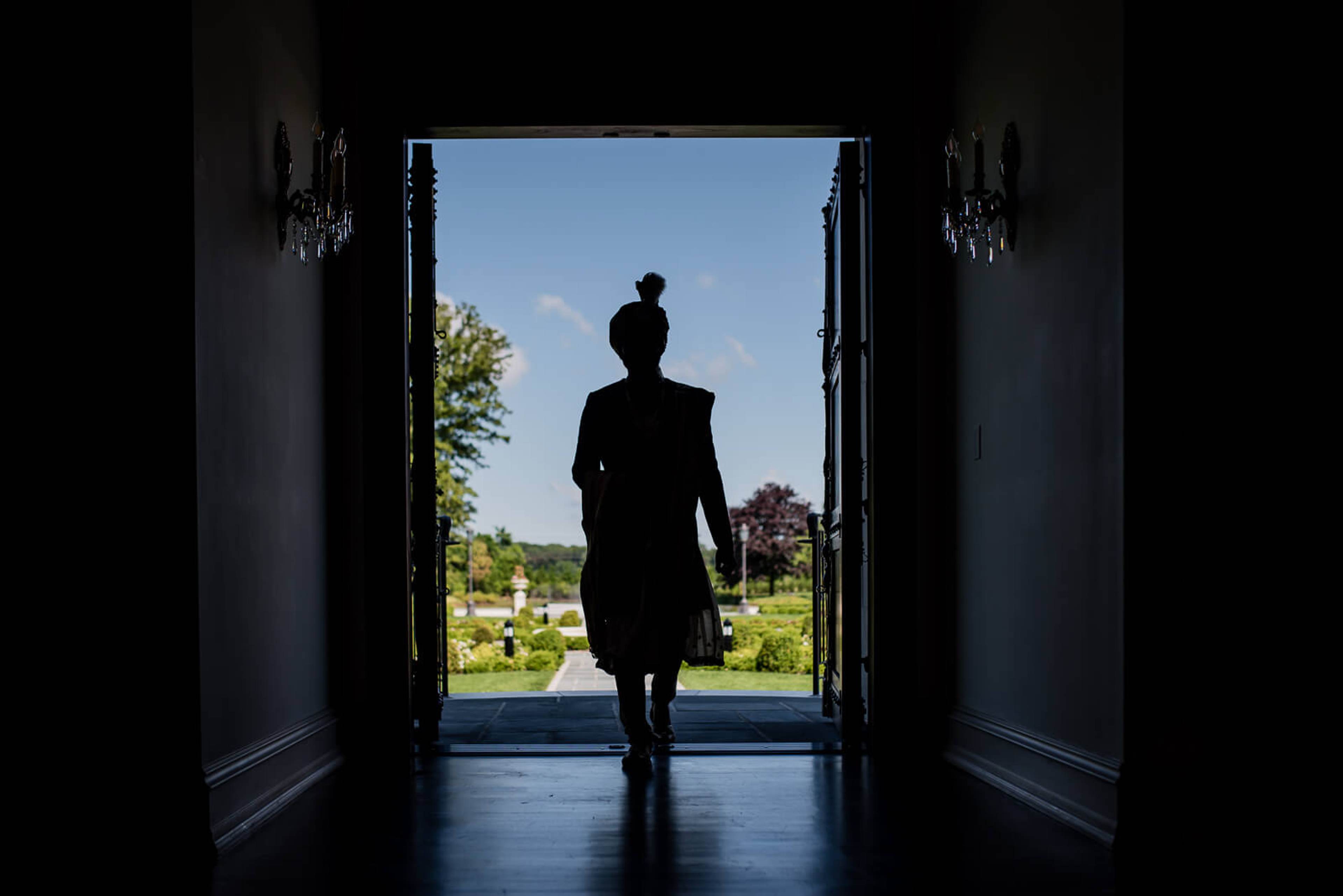 Silhouetted figure in dark dress walking through open doors toward sunny garden landscape