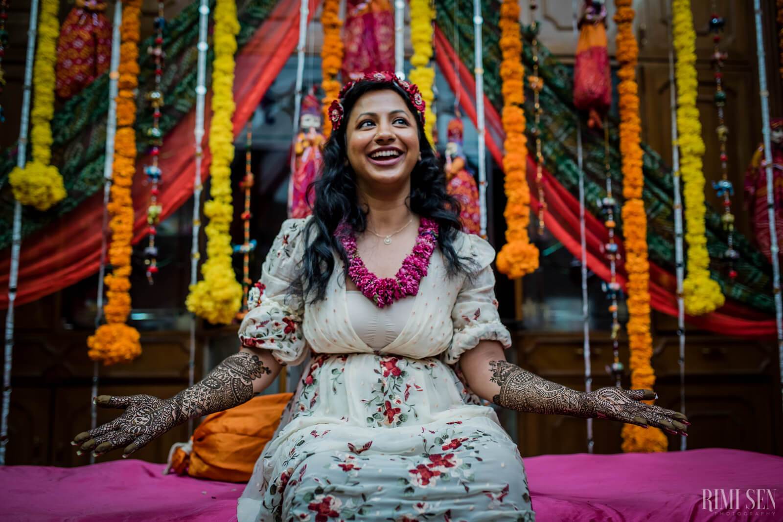 Nivetha smiling in floral dress with henna, vibrant marigold and red fabric garland backdrop