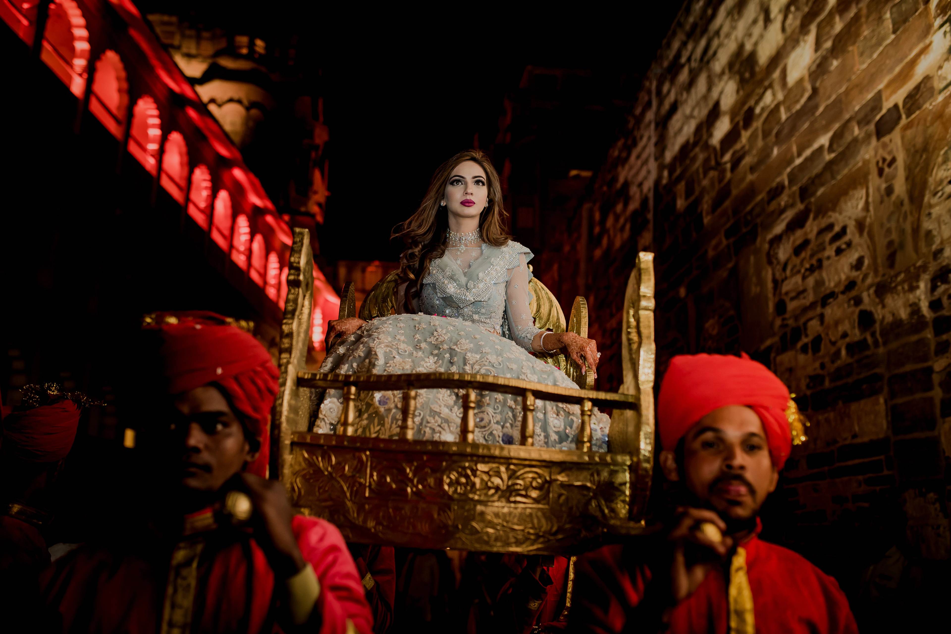 Woman in ornate lace dress seated on golden chair, two men in red caps and traditional dress beside her, dramatic red lighting