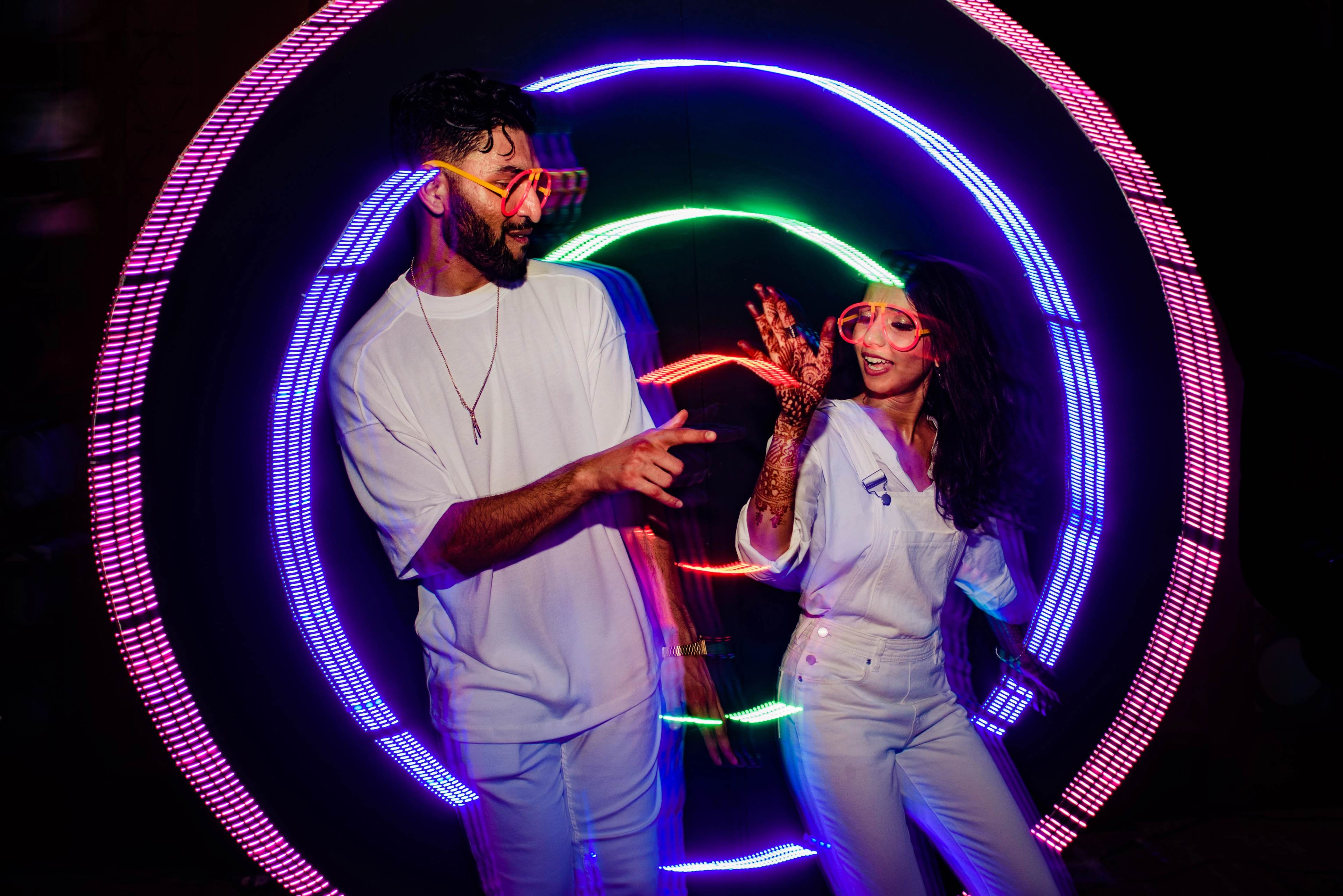 Two people dancing inside colorful neon light rings, wearing white casual attire and smiling