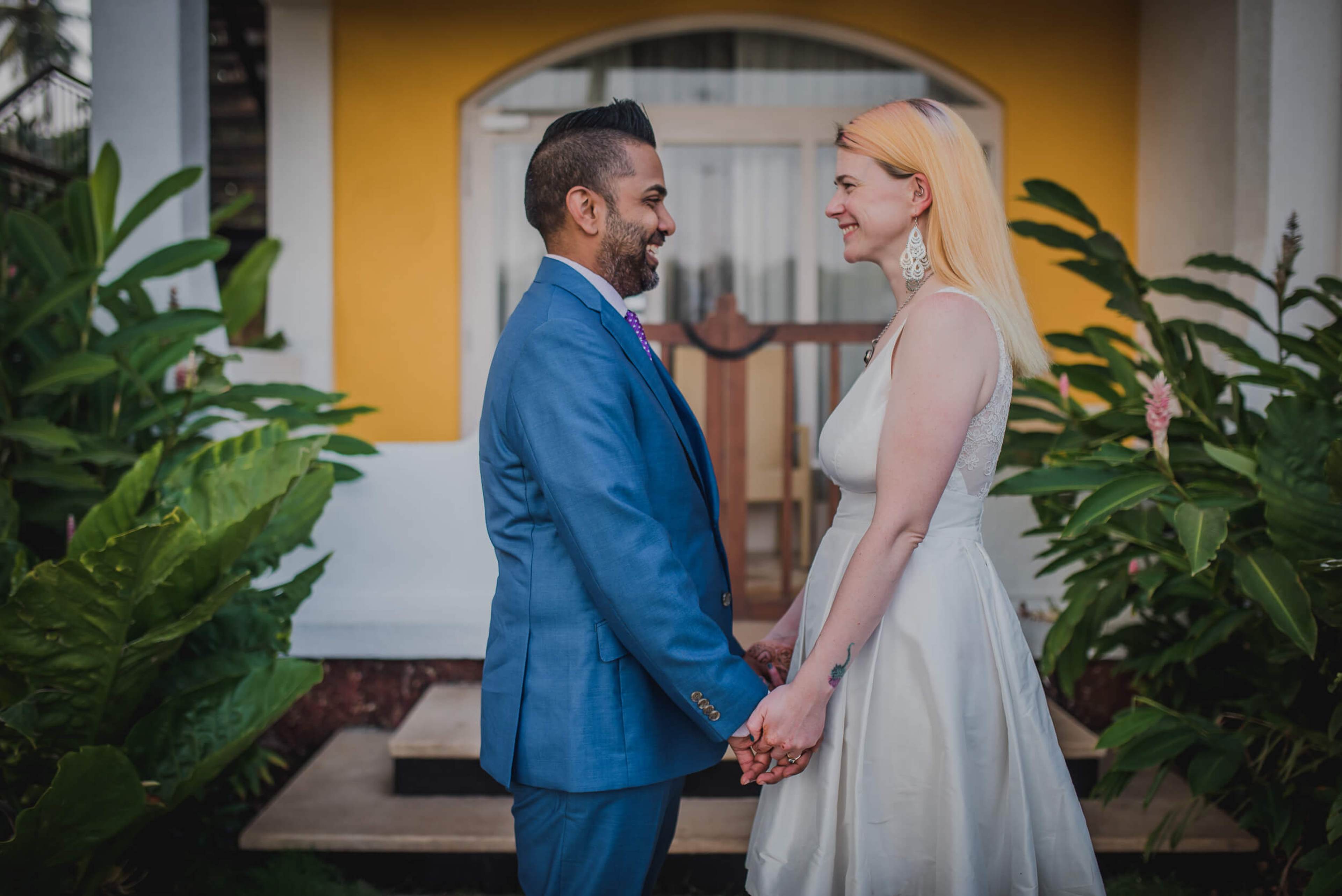 Sarah and Ravin hold hands amid lush plants, smiling at each other in Goa's yellow-walled courtyard