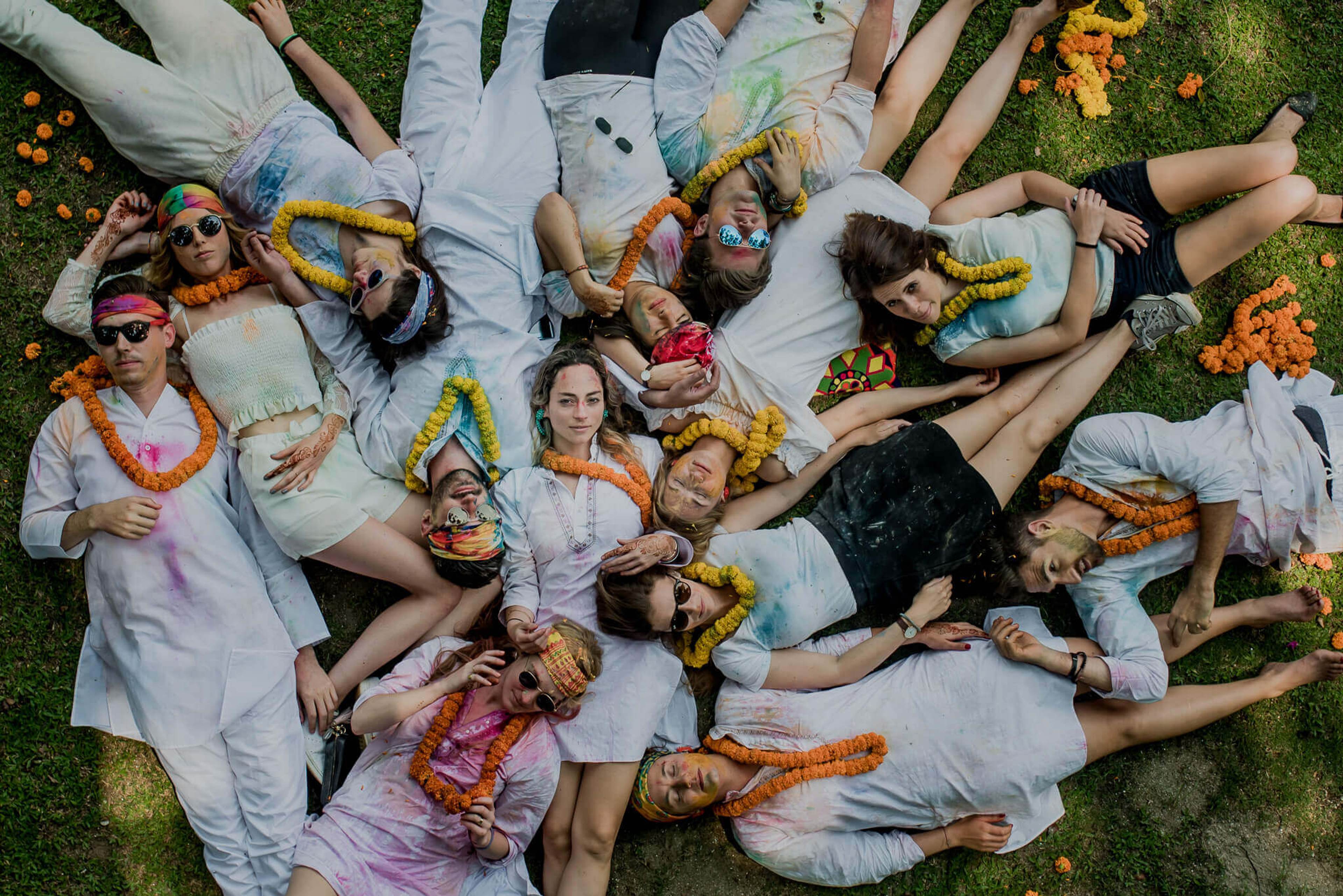 Group of friends lying on grass in white clothing with yellow and orange flower garlands, overhead view