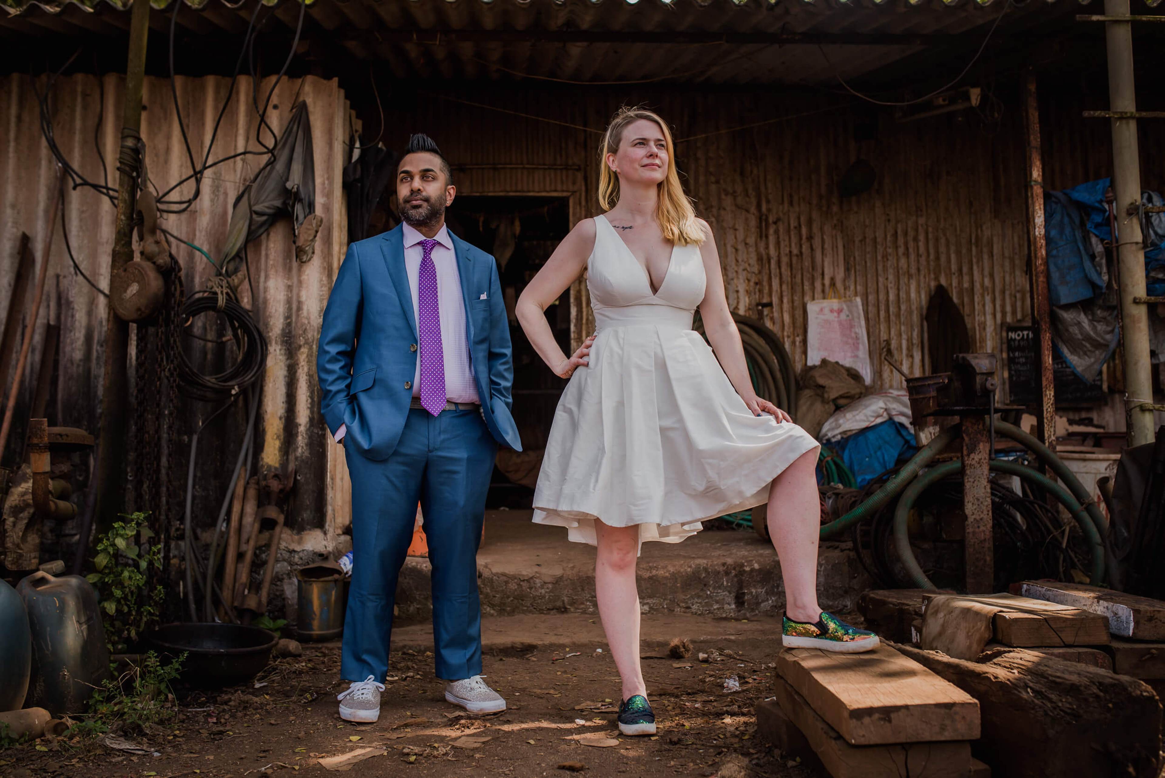 Sarah and Ravin stand in a rustic Goa barn, man in blue suit, woman in white dress, surrounded by vintage machinery