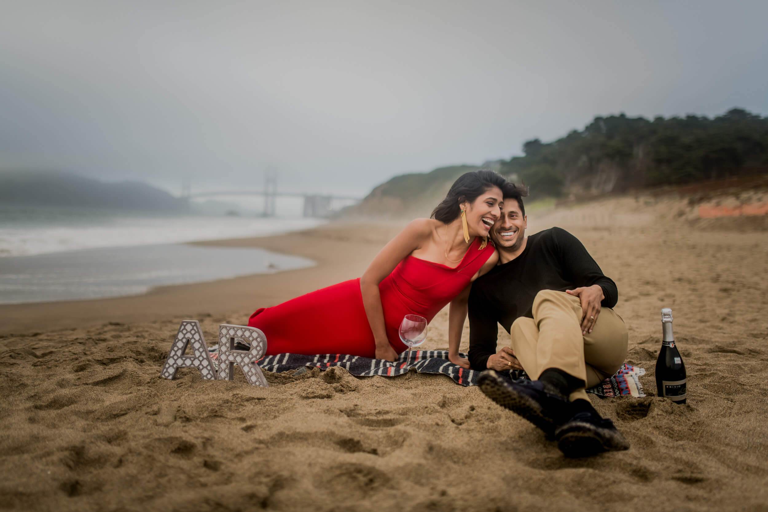 Anu and Raghu sit together on sandy California beach in red dress and black shirt, Golden Gate Bridge visible in misty distance