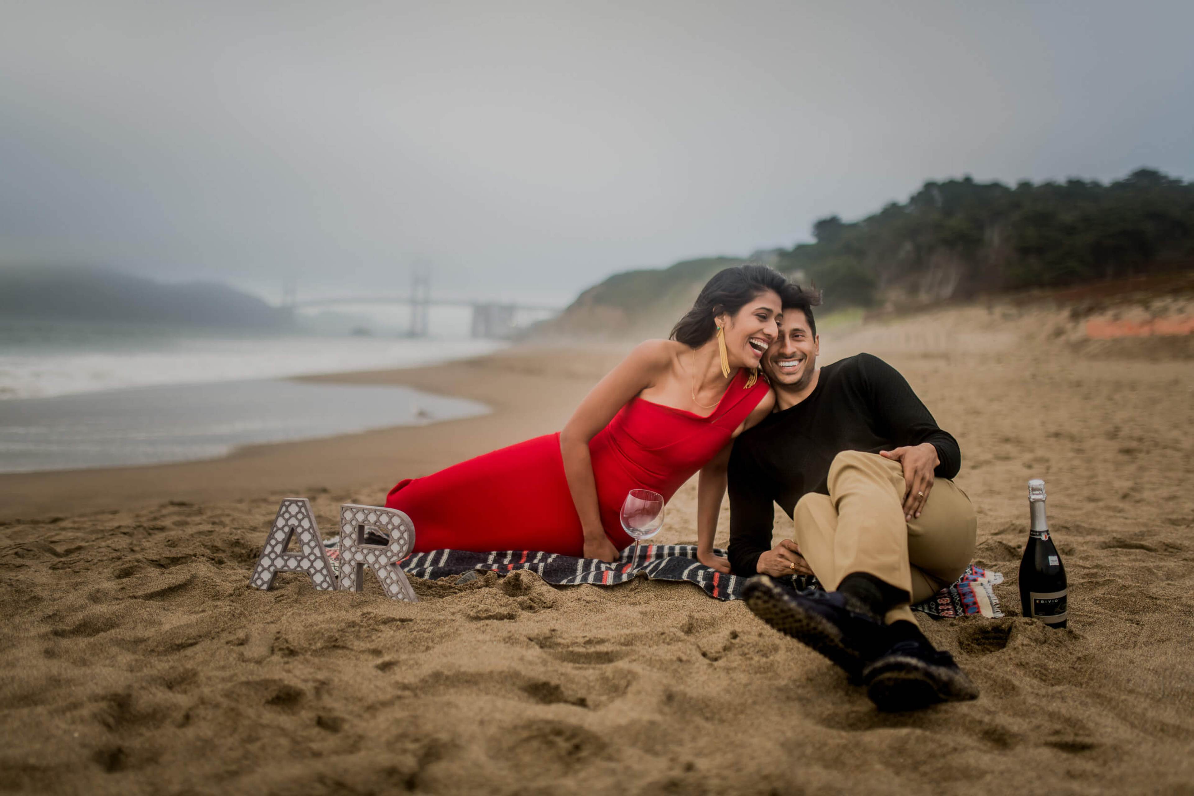 Couple in red dress and black jacket sit together on misty beach sand with champagne and blanket