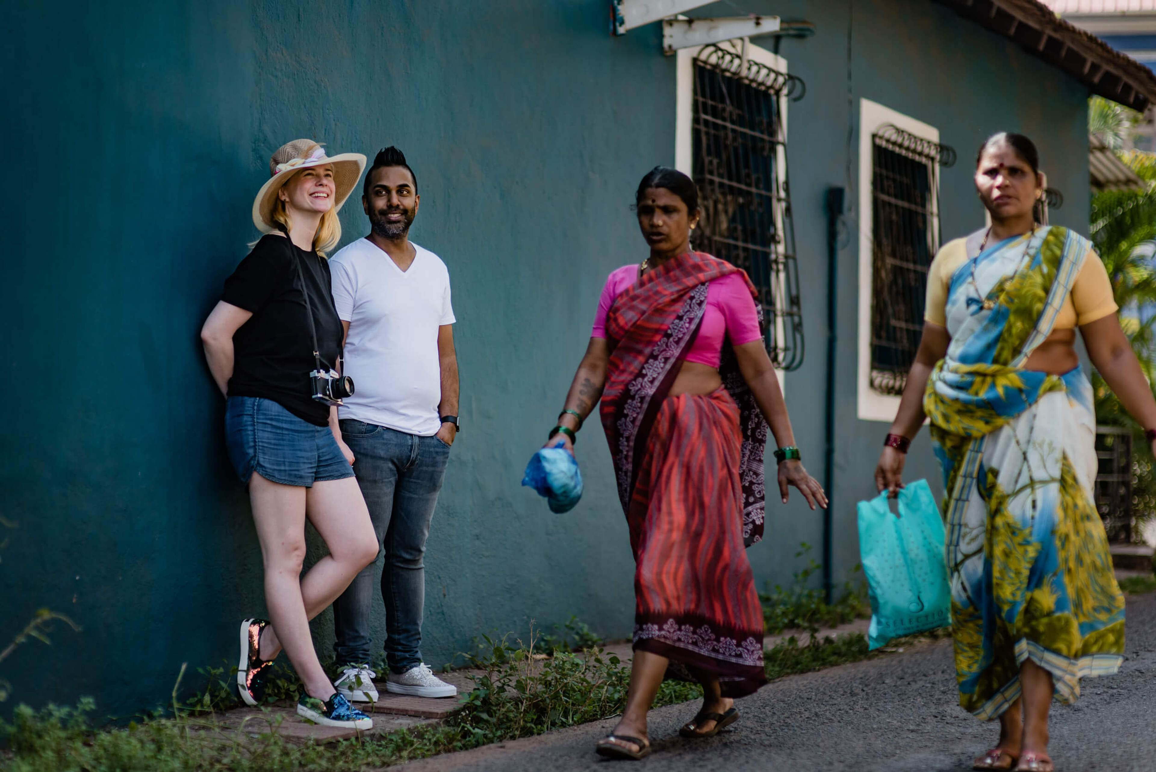 Sarah and Ravin with local women against vibrant teal building wall in Goa