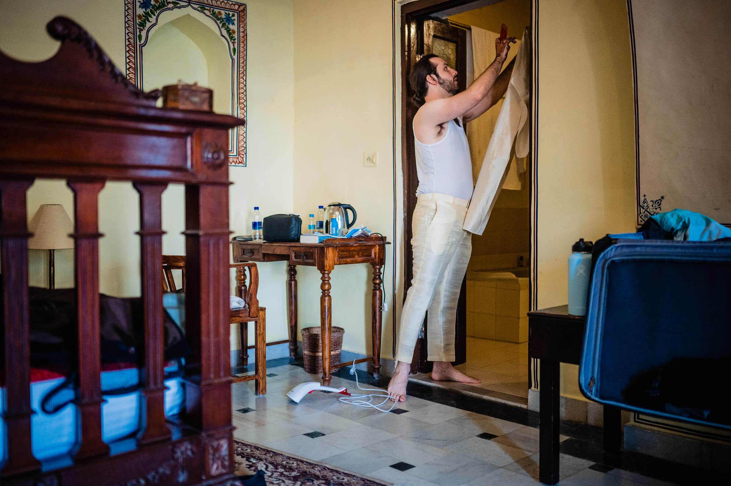 Maitrayee adjusting wedding attire in ornate Rajasthan room with traditional furniture and blue luggage