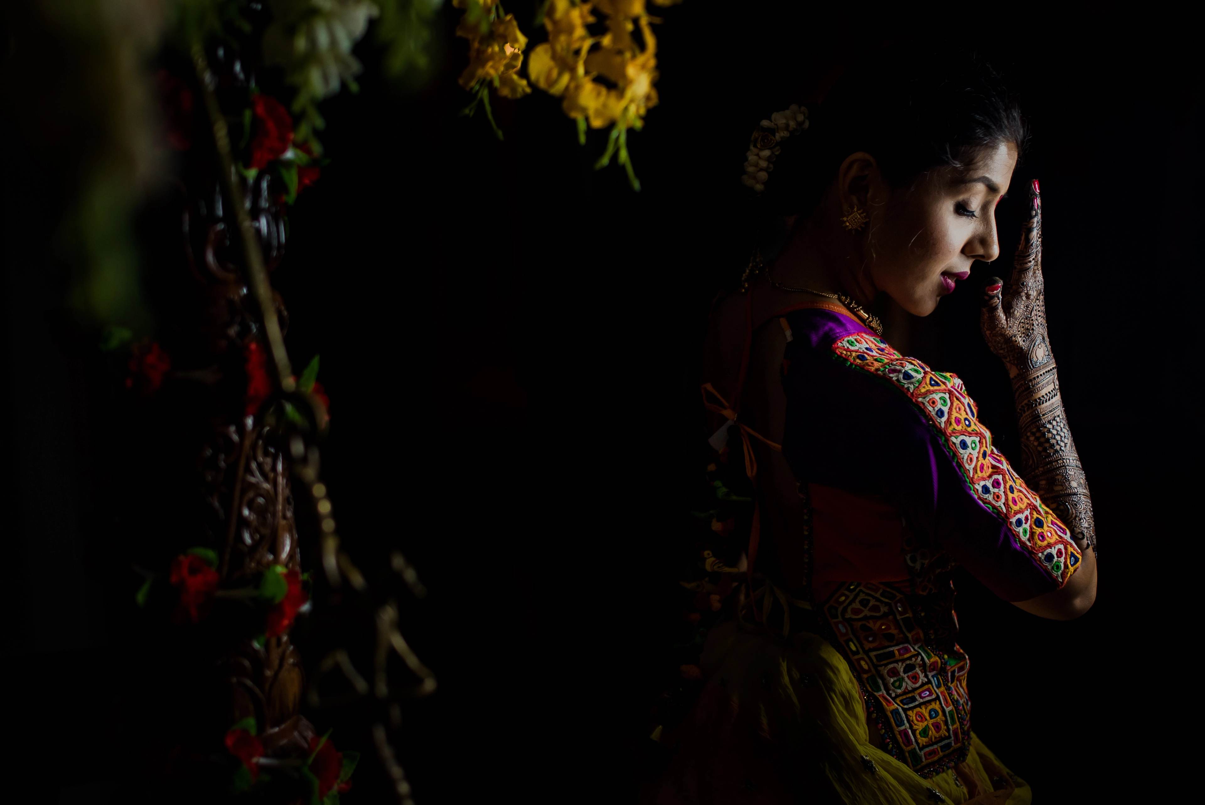 Woman in ornate embroidered traditional attire gazes downward, framed by hanging flowers against dark background