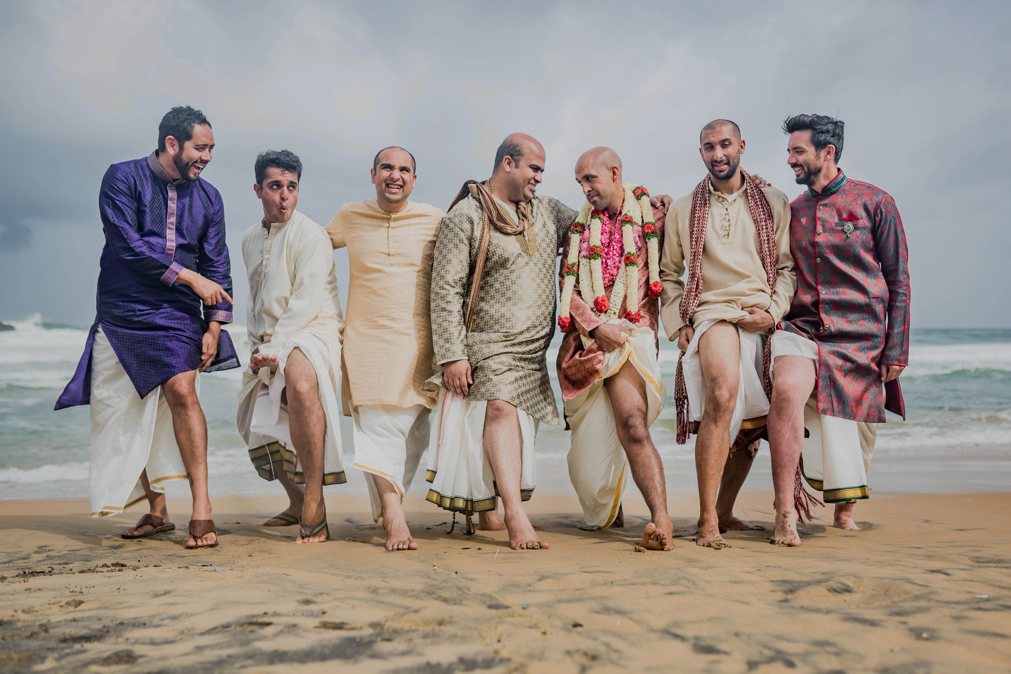 Seven men in traditional South Asian attire walking barefoot on a beach together