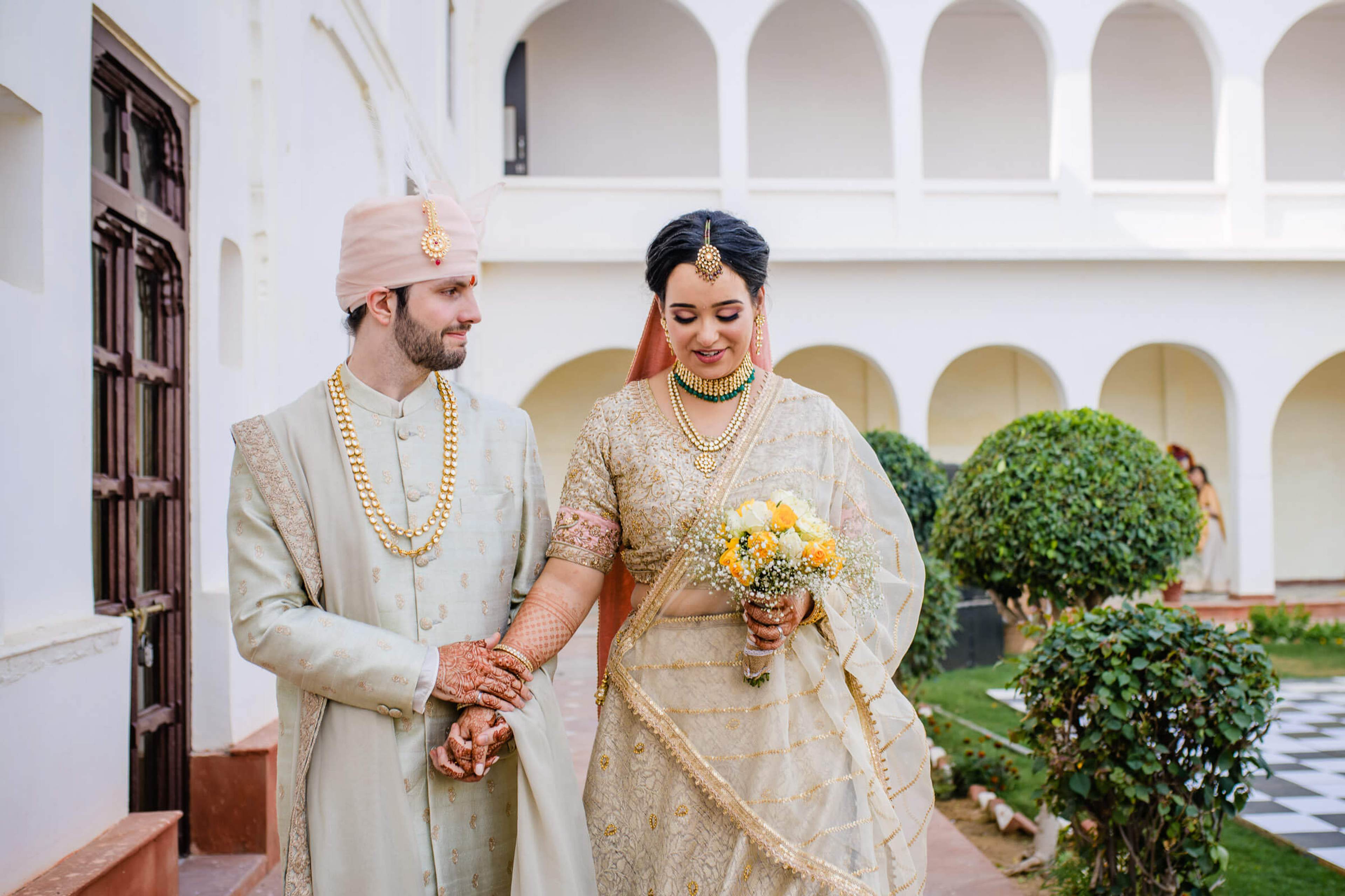 Maitrayee and Eric hold hands in traditional Indian wedding attire within a white-arched Rajasthan courtyard