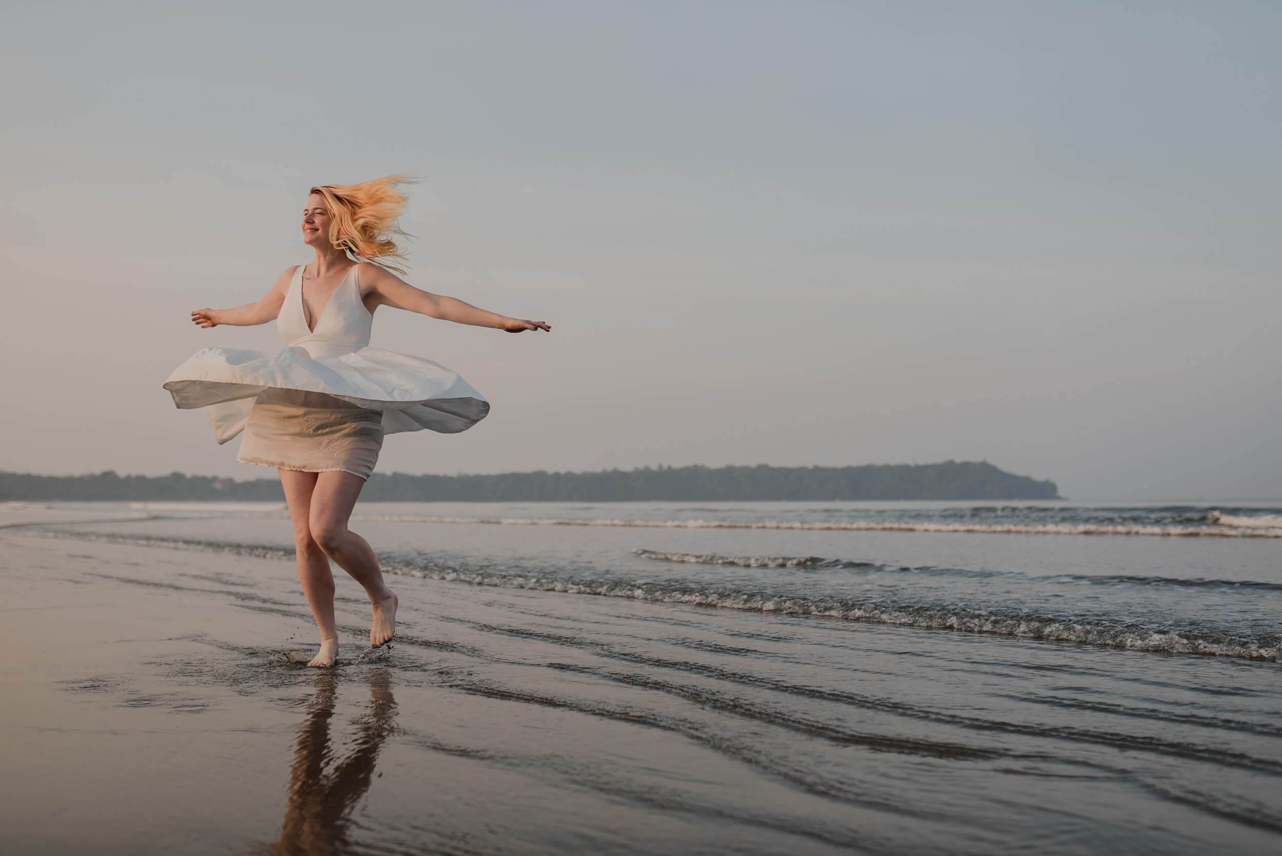 Sarah twirling in white dress on wet Goa beach at sunset, arms outstretched, wind-swept hair