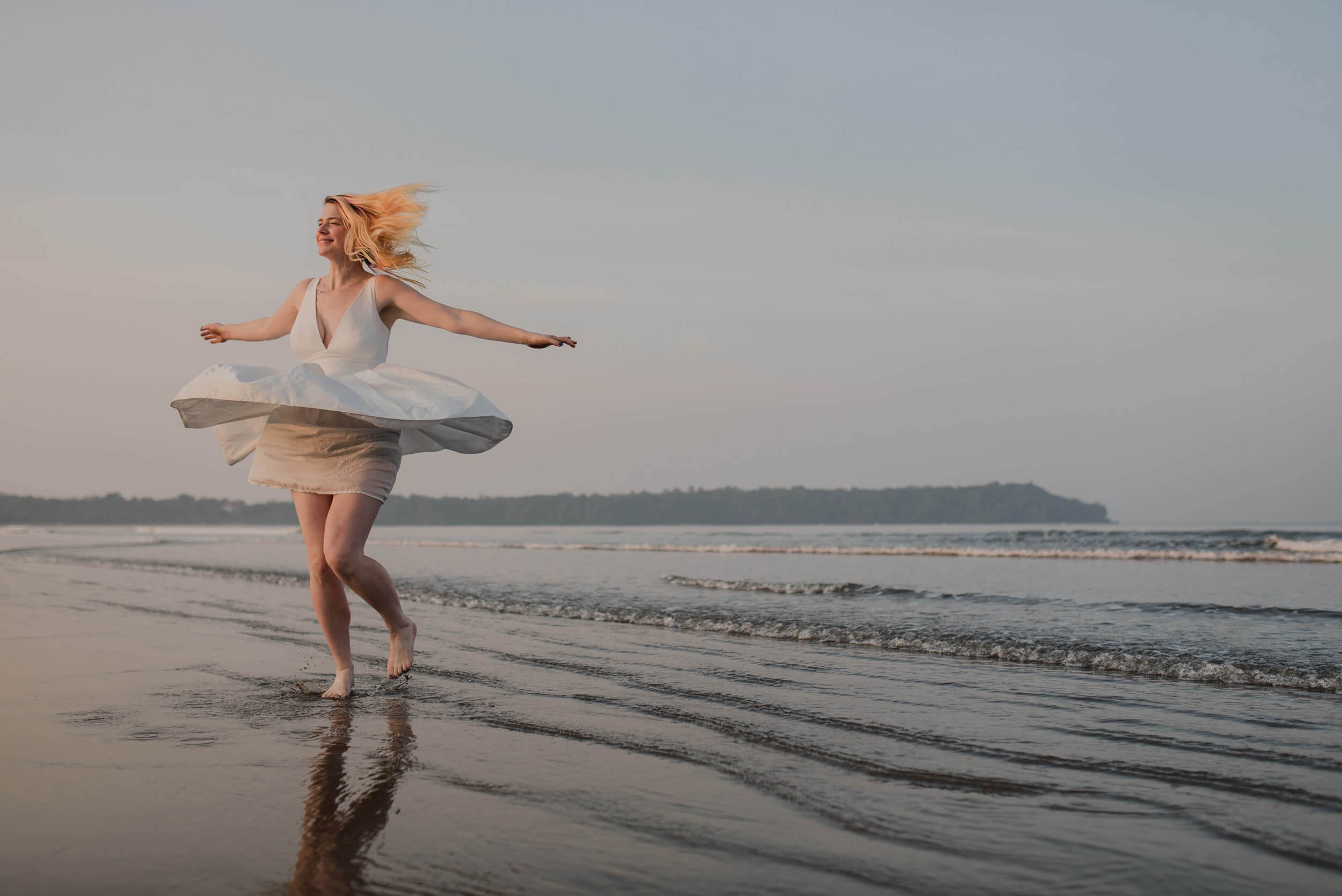 Sarah twirling in white dress on wet Goa beach at sunset, arms outstretched, wind-swept hair