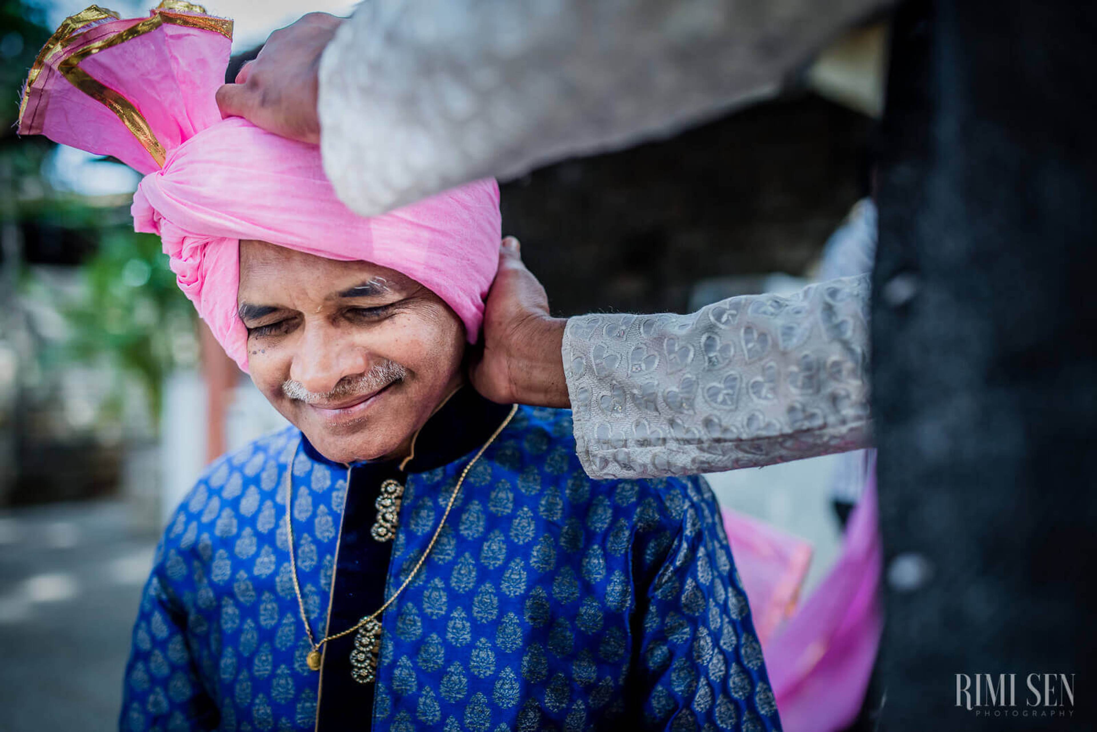 Groom Aniket in blue patterned sherwani and pink turban, smiling as hand adjusts his headwear