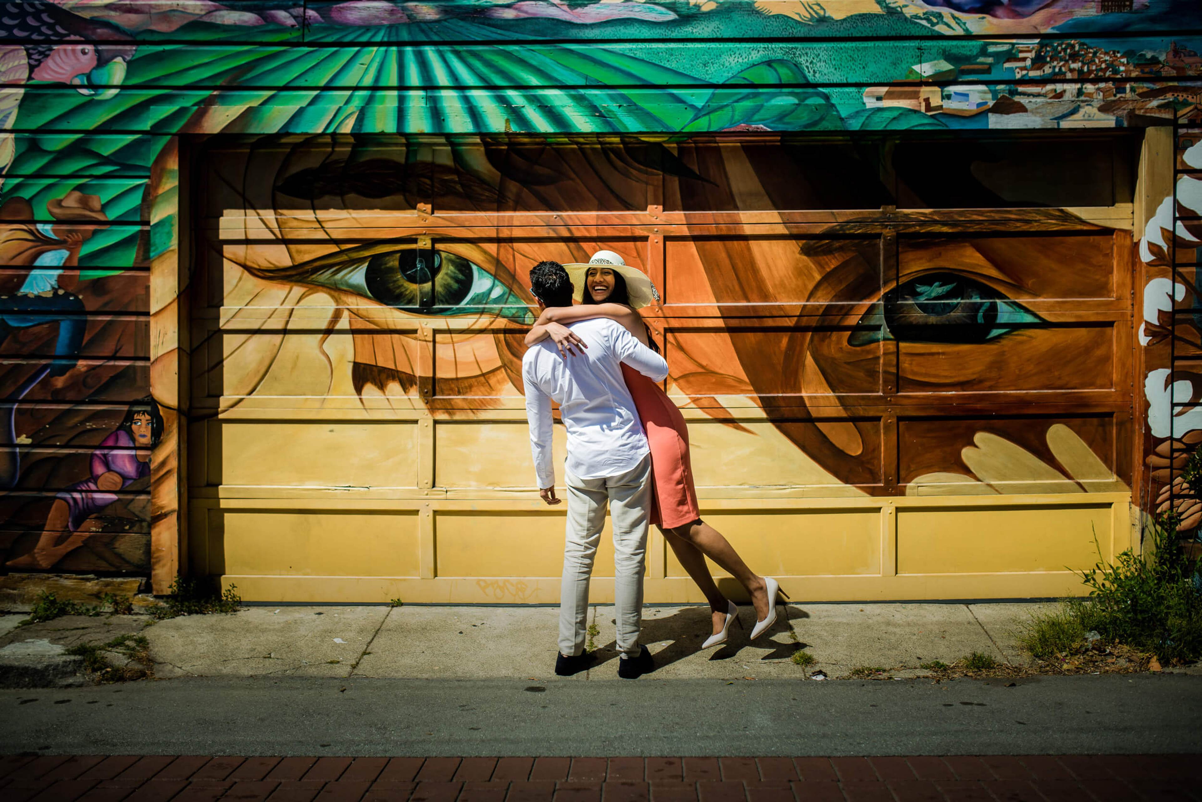 Anu and Raghu embrace against vibrant street mural in California, dappled sunlight casting shadows