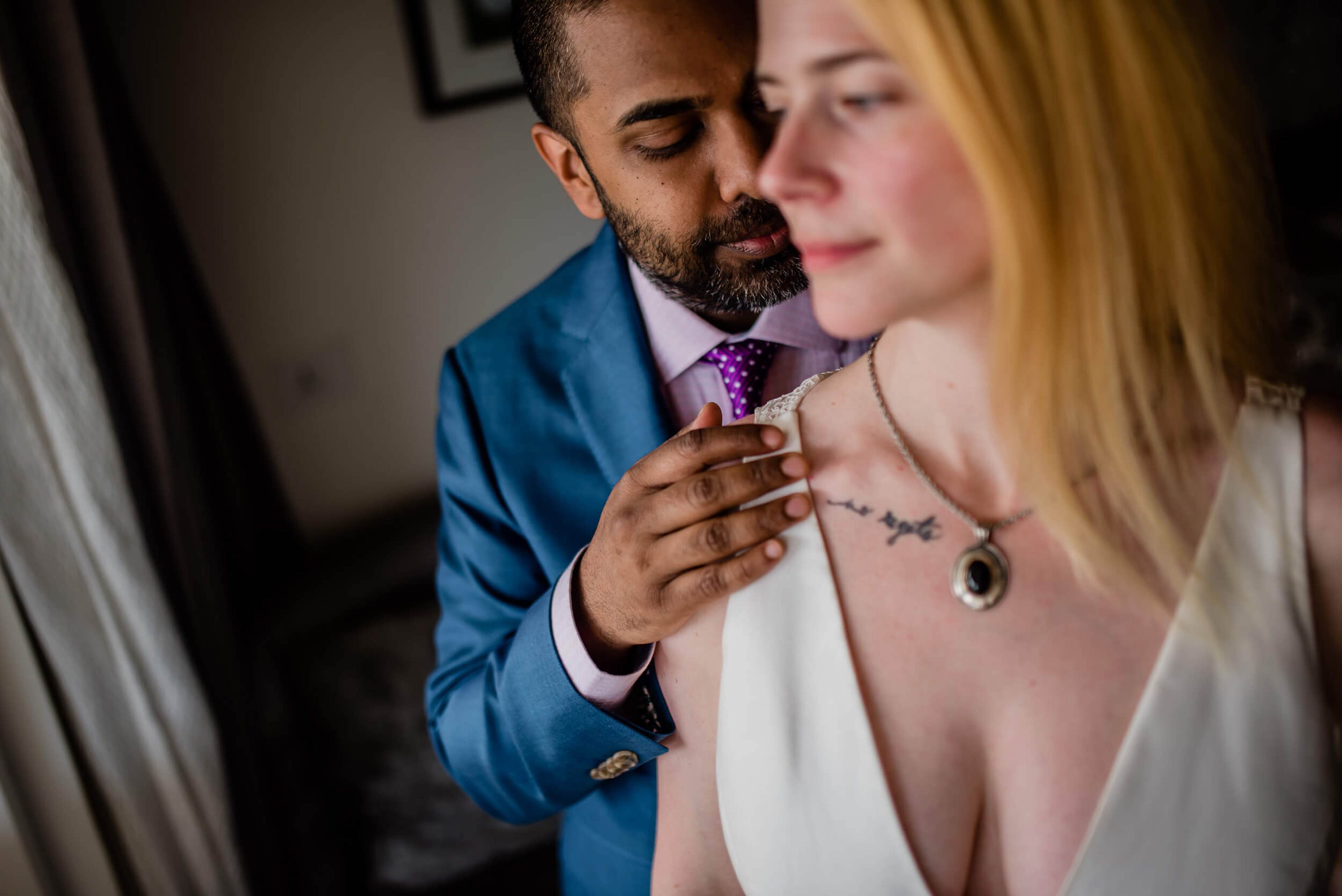 Sarah and Ravin embrace in soft indoor light, he in blue suit and purple tie, she in white with delicate necklace